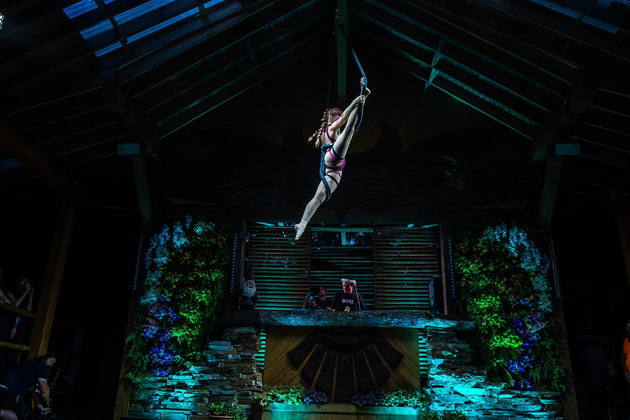 A woman in a striped dress is performing aerial acrobatics on a hoop hanging from the ceiling in a decorated indoor venue with wooden beams and brick wall. There are audience members watching.