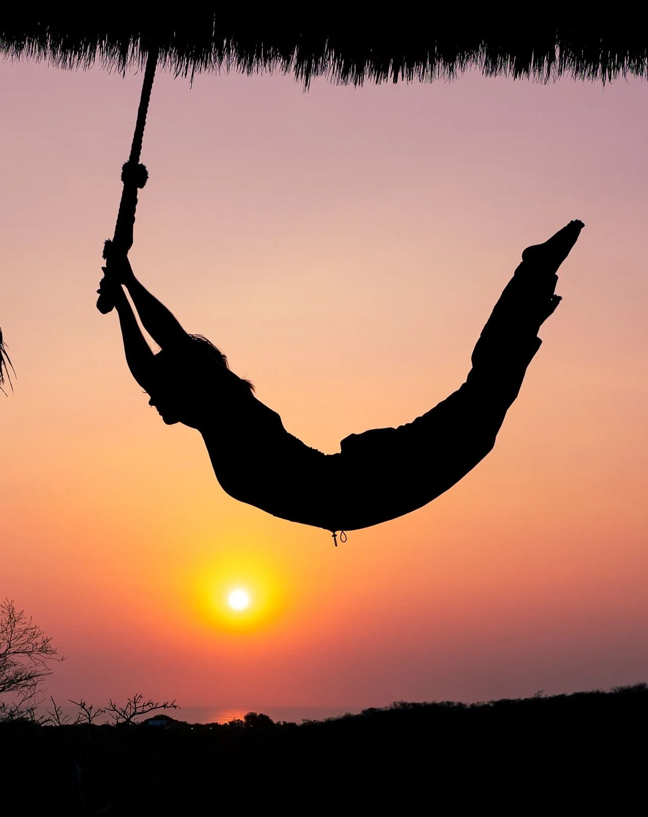 Silhouette of a person on a hammock hanging from a thatched roof, with the sunset sky in the background.