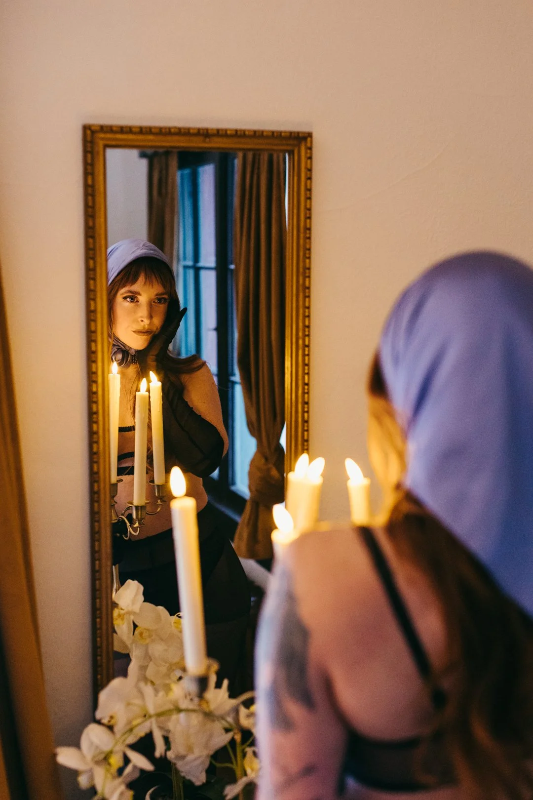 A woman with a purple headscarf and dark tattoo on her arm looking at her reflection in a mirror surrounded by lit candles and white flowers.