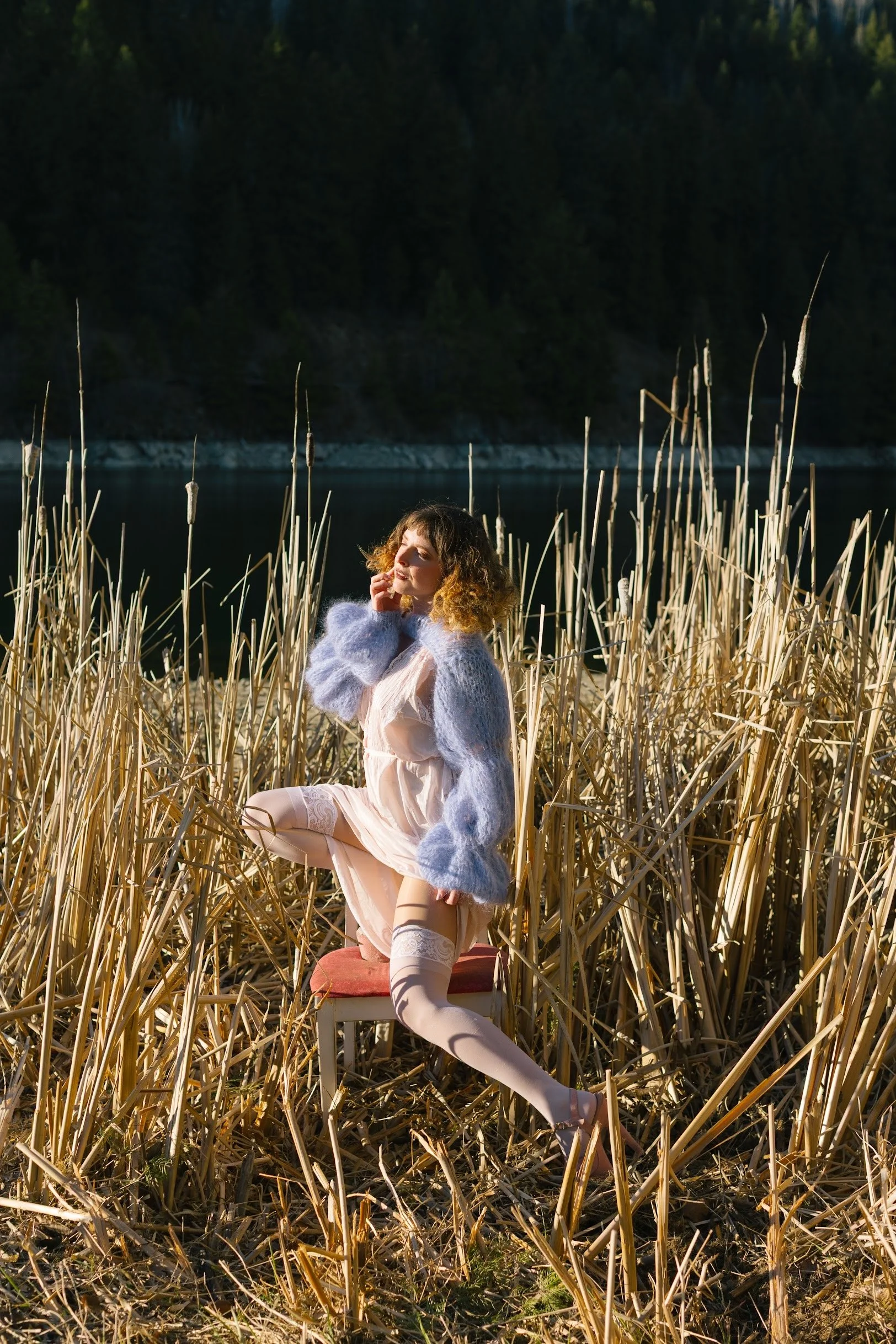 A woman with curly hair sitting on a chair in a field of tall, dry reeds by a lake with a forested hillside in the background, dressed in pastel pink lingerie, stockings, and a light blue fuzzy sweater.