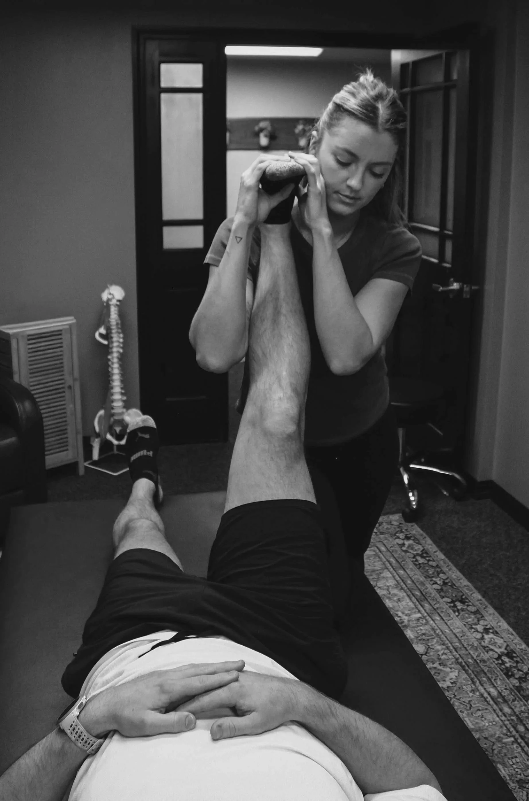 A woman is performing a physical therapy or chiropractic adjustment on a man's leg in a room with a patterned rug and a door in the background.