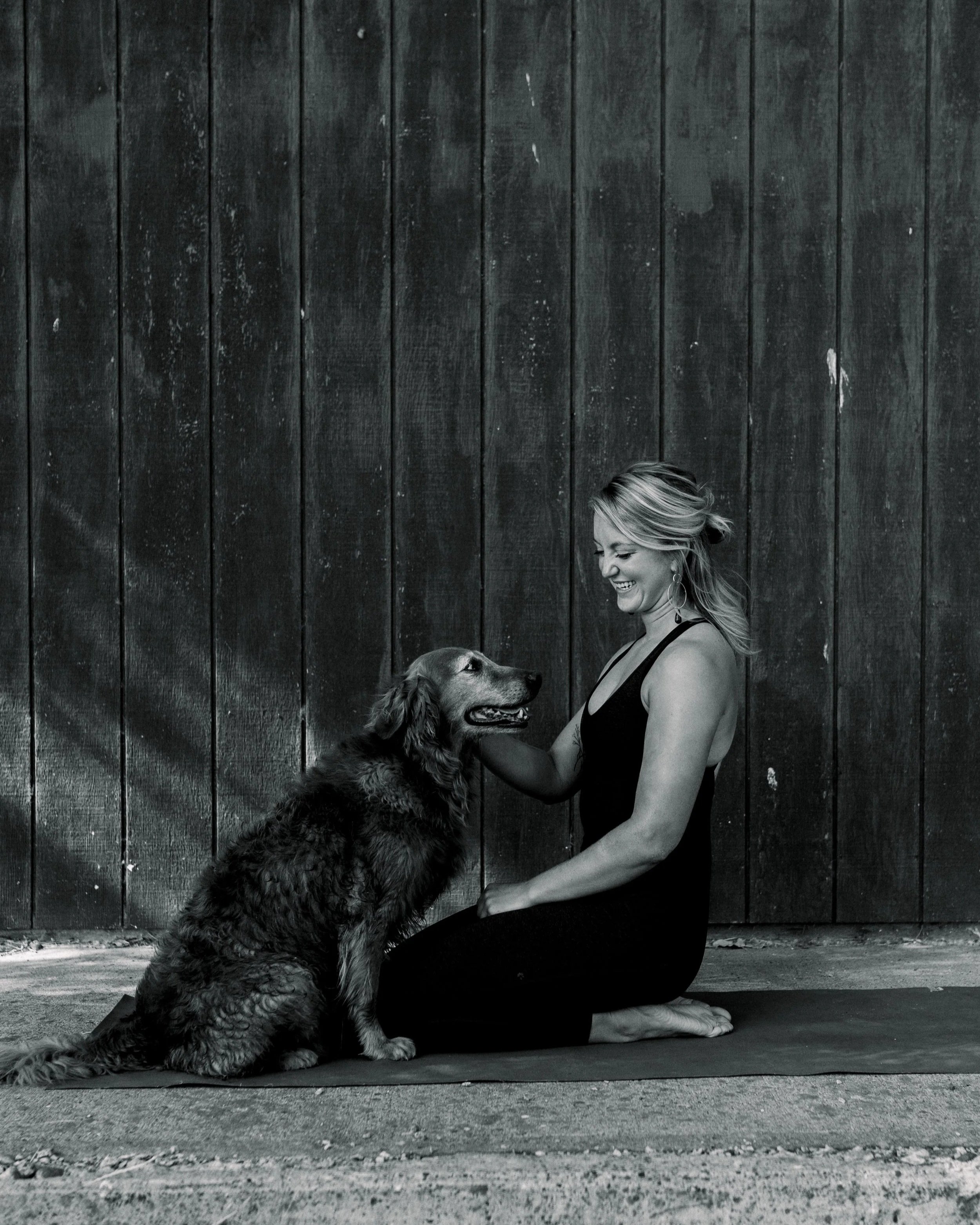 Black and white photo of a woman practicing yoga outdoors with her dog, sitting on a yoga mat against a wooden fence.