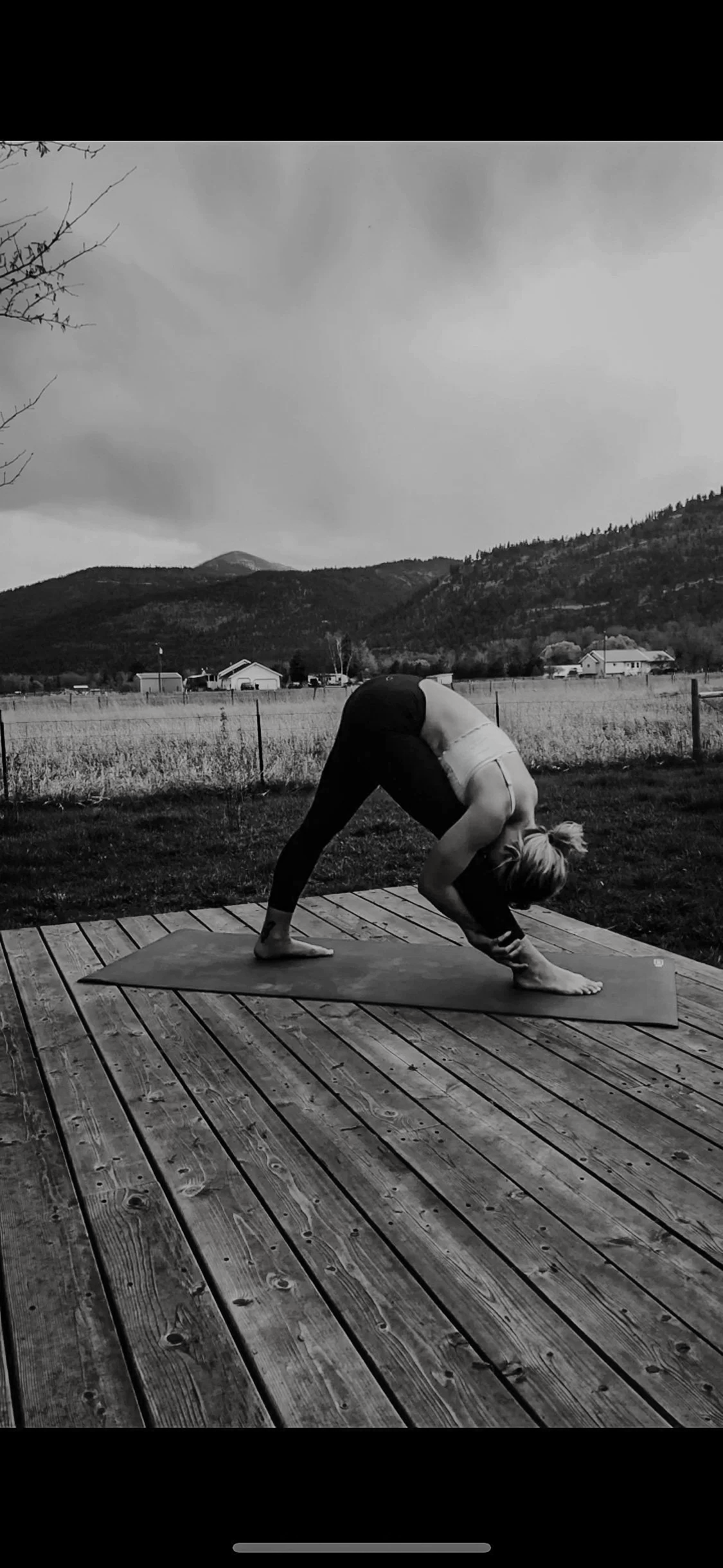 A person practicing yoga outdoors on a wooden deck, performing a forward bend pose with their hands on the ground and feet apart, overlooking a rural landscape with hills, houses, and cloudy sky.