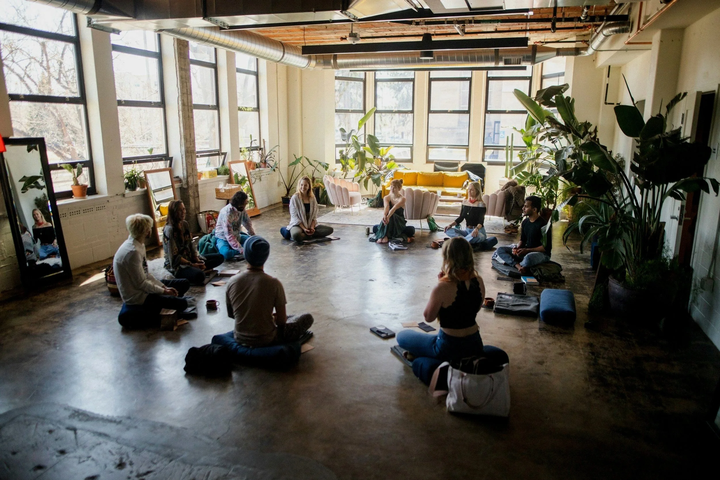 Group of people sitting on the floor in a circle during a meeting or workshop in a spacious, well-lit room with large windows and indoor plants.