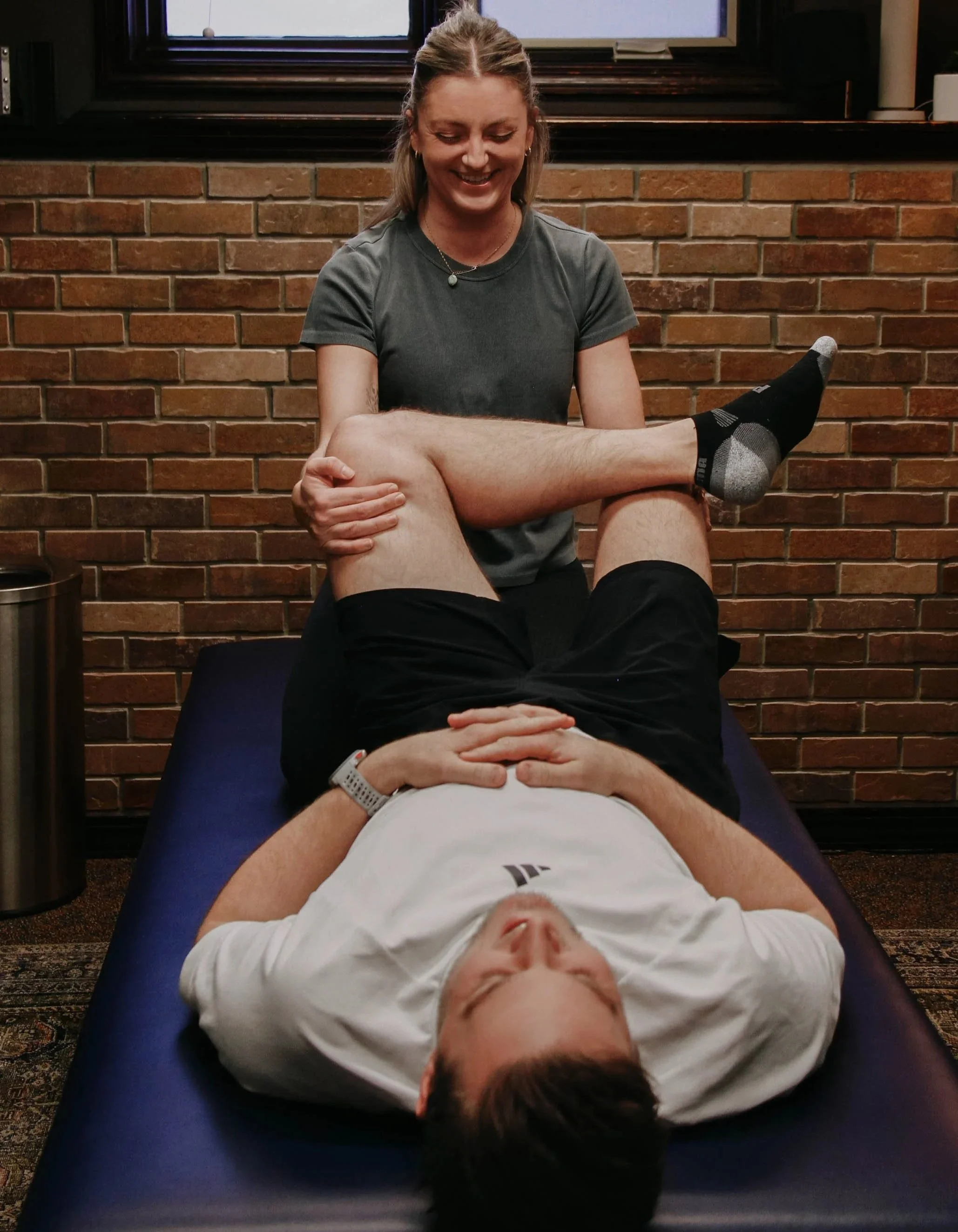 A woman with blonde hair tied back smiling and sitting behind a man lying on his back on a massage table, holding his leg. The man has brown hair, is dressed in a white shirt and shorts, and is wearing a hospital wristband. They are in a room with a brick wall and a window.
