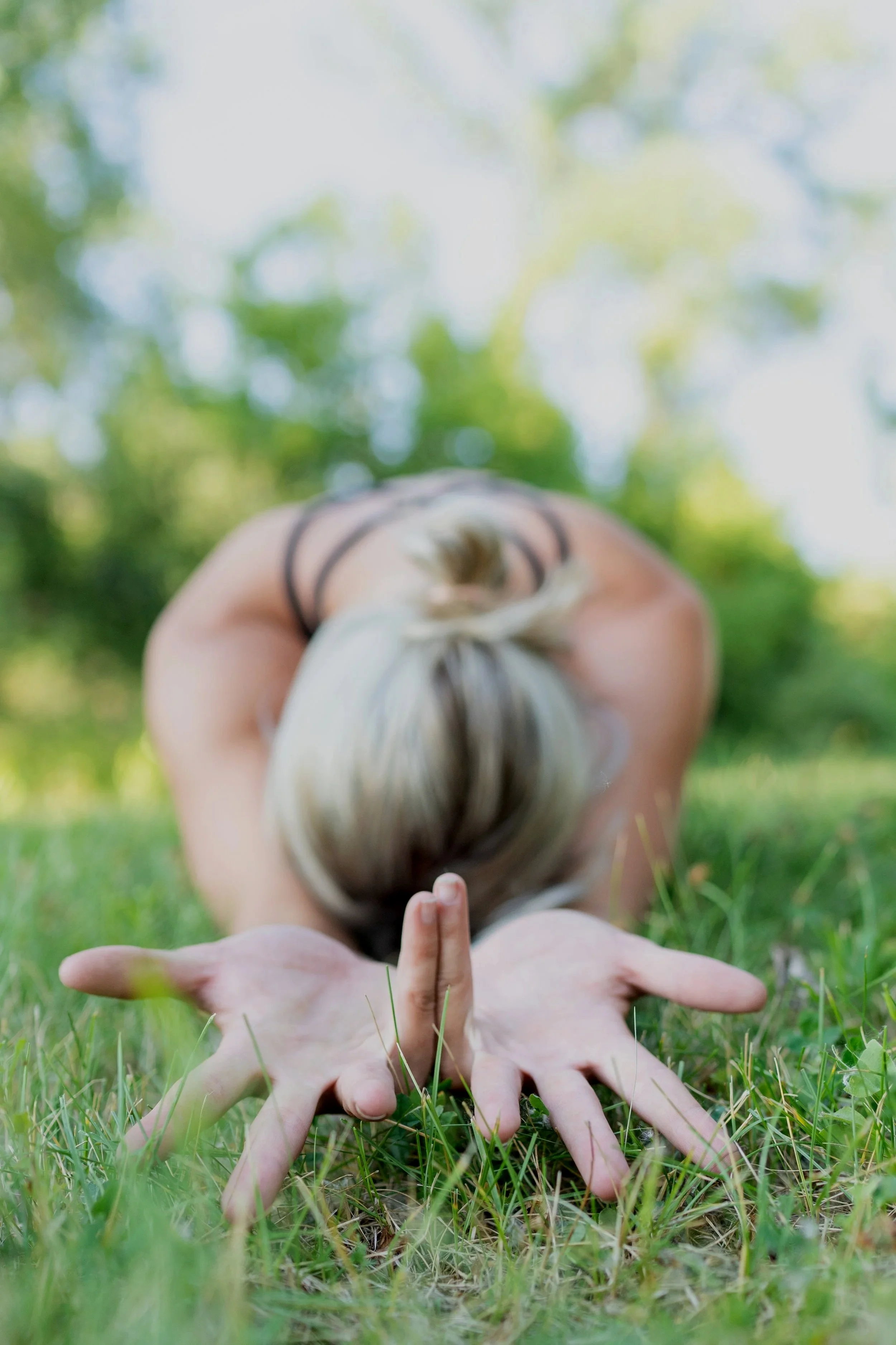 Person practicing yoga outdoors on grass, in a forward bend with hands on the ground, head down, surrounded by greenery.