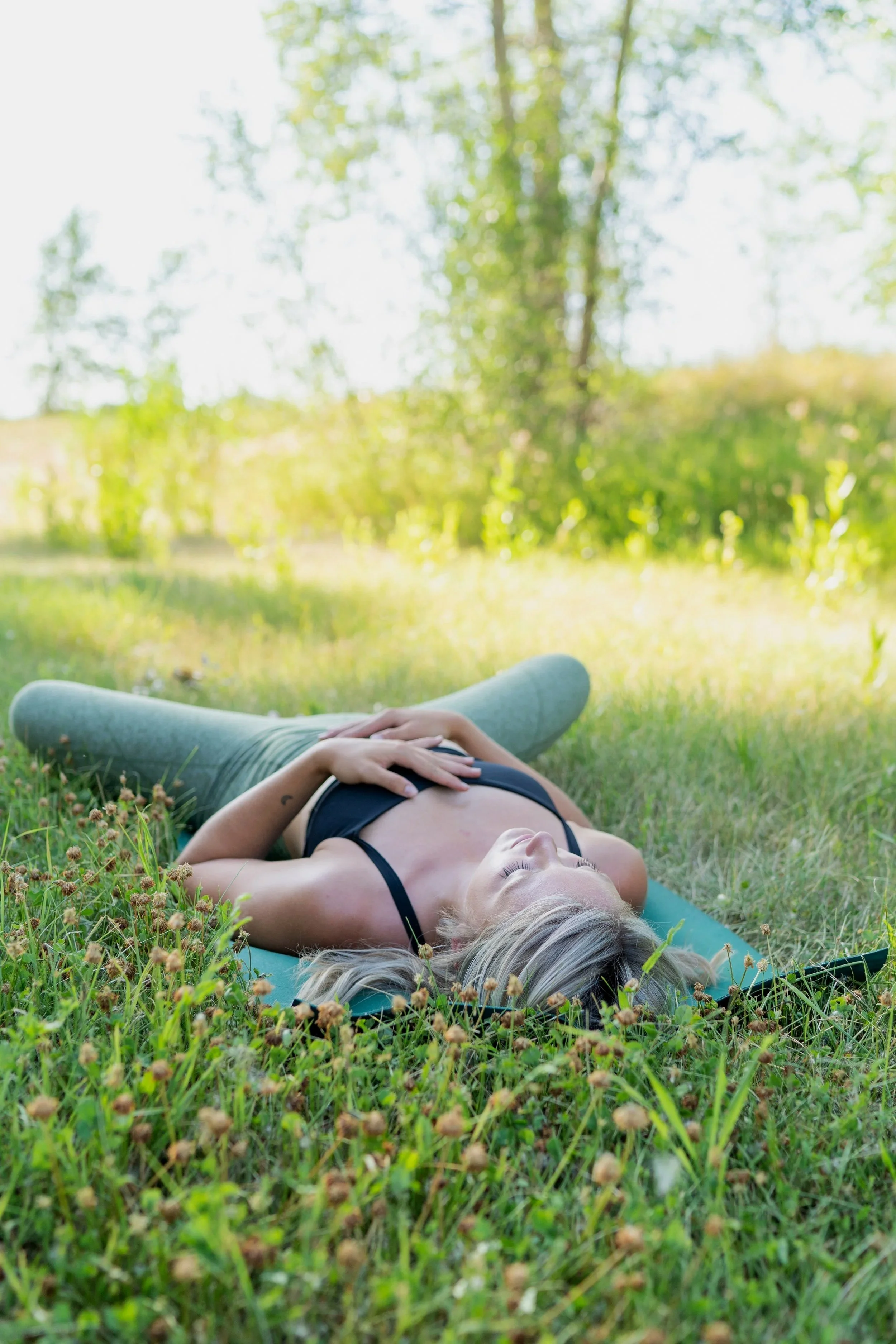 A woman with blonde hair in a black top laying on her back on a blue yoga mat in a grassy field, with her eyes closed and hands resting on her abdomen, surrounded by greenery and trees in the background.