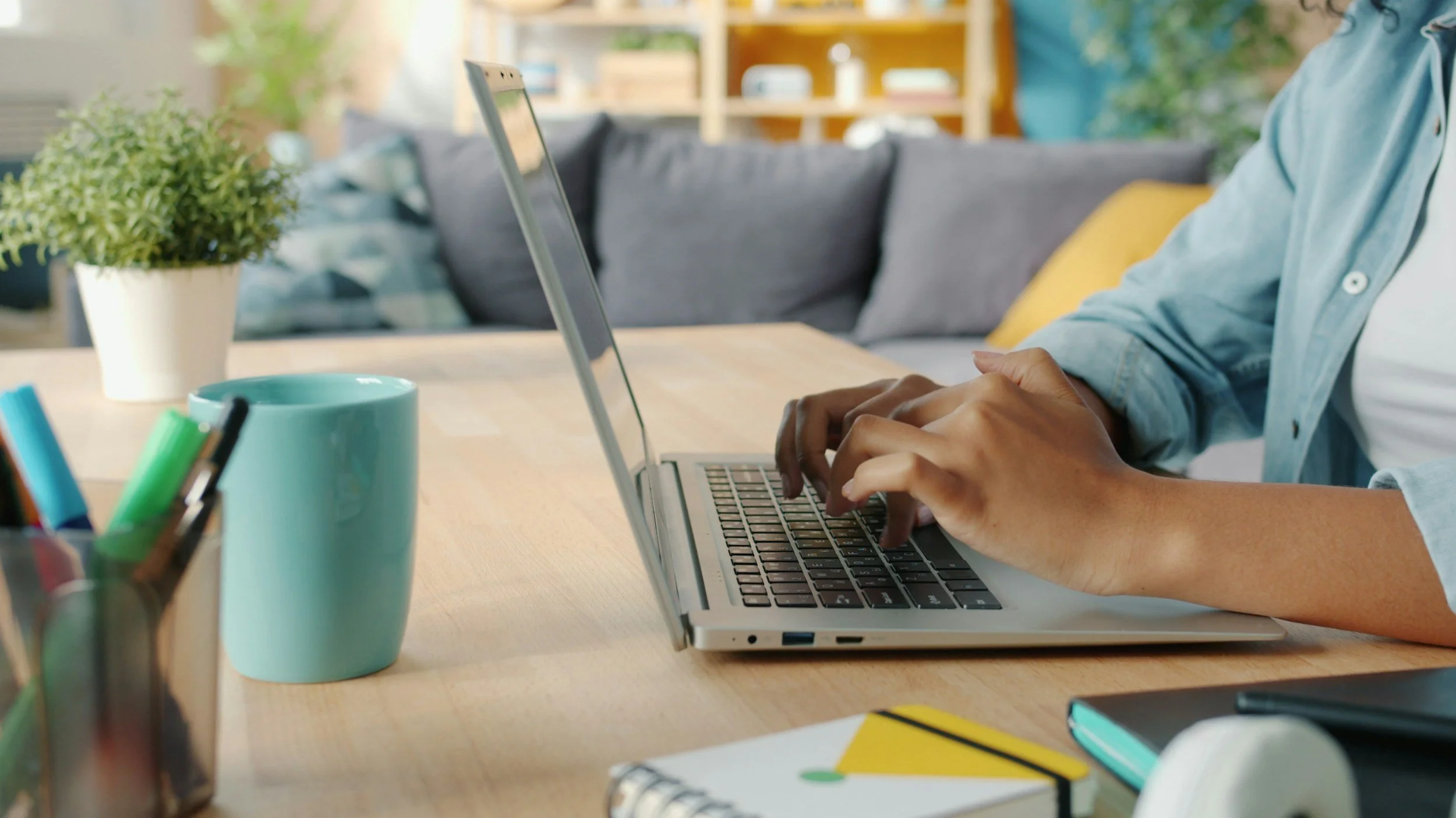 Person typing on a laptop at a wooden desk in a room with bookshelves and houseplants.