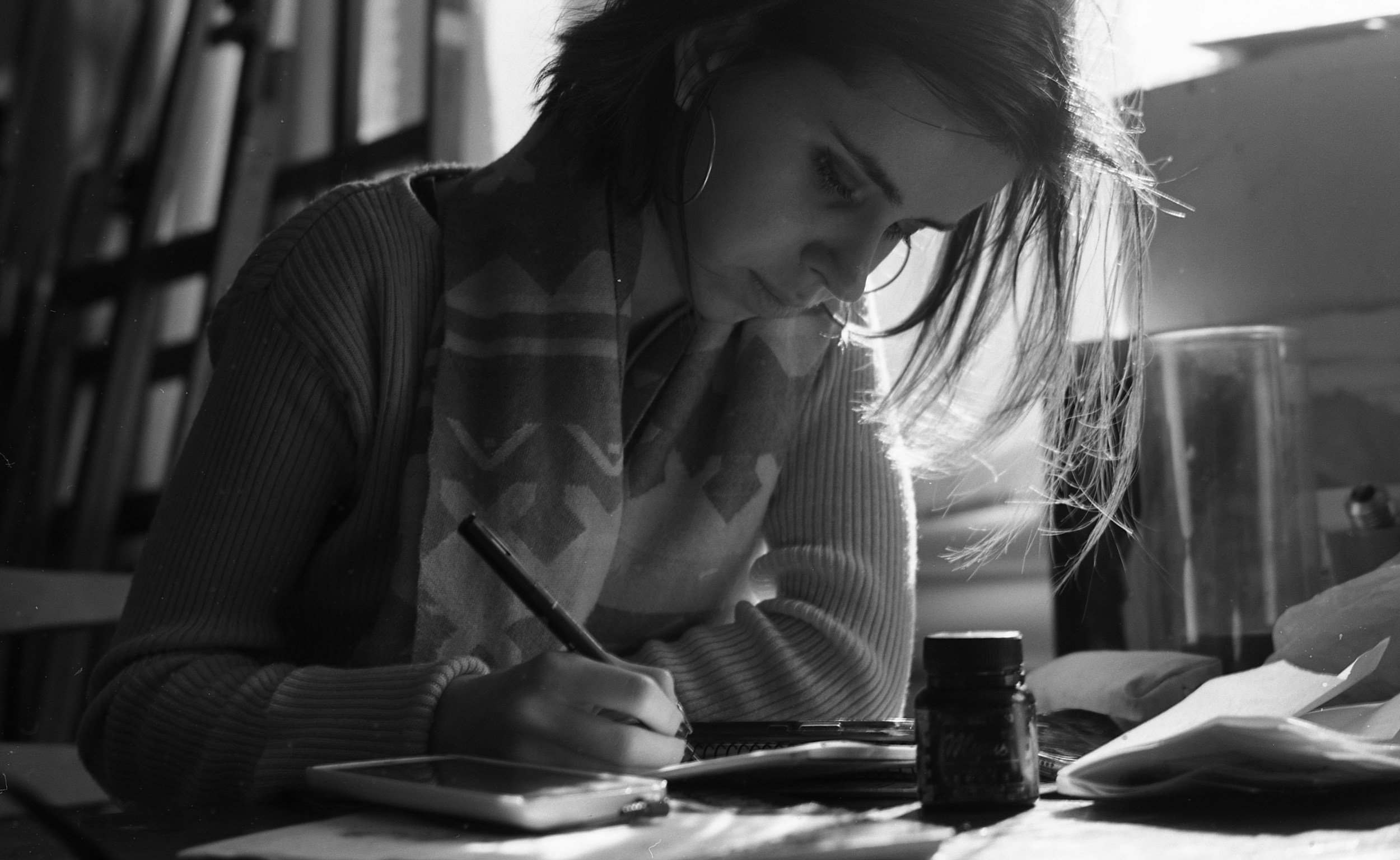 A woman with short hair, glasses, and hoop earrings is sitting at a table indoors, writing or drawing on paper with a pen. The table has a bottle, and behind her, there are shelves with books.