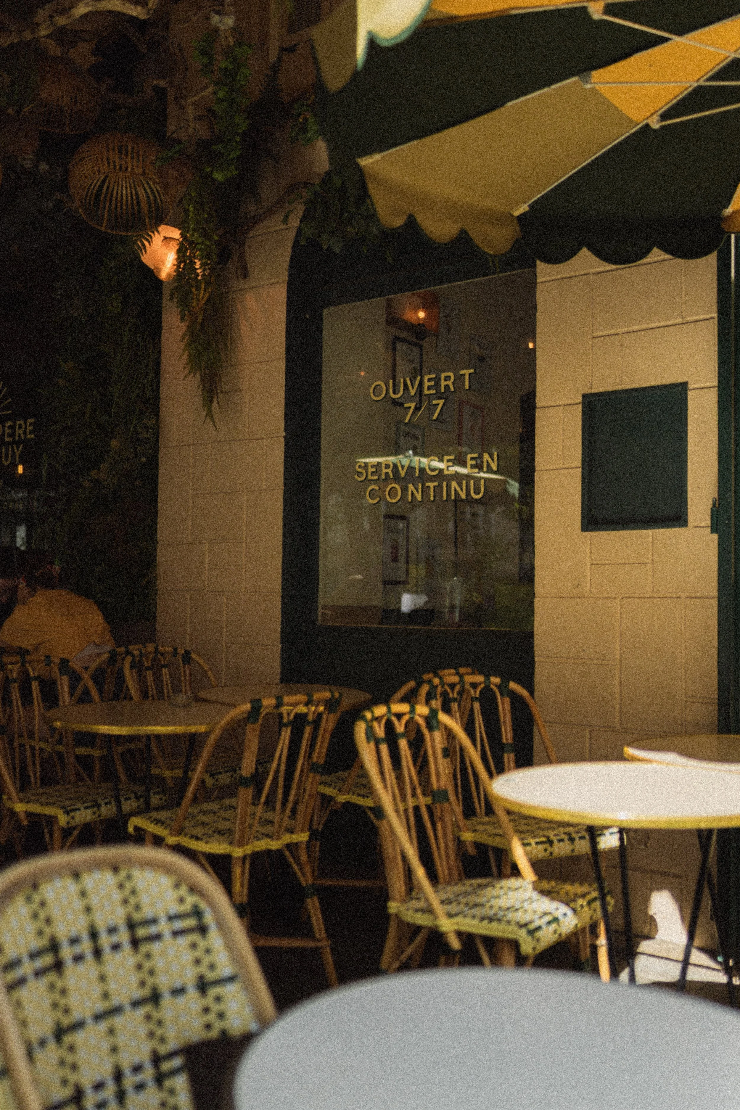 Night view of outdoor seating area with tables and chairs, a window with French words 'OUVERT 7/7 SERVICE EN CONTINU', and a yellow and green striped awning.