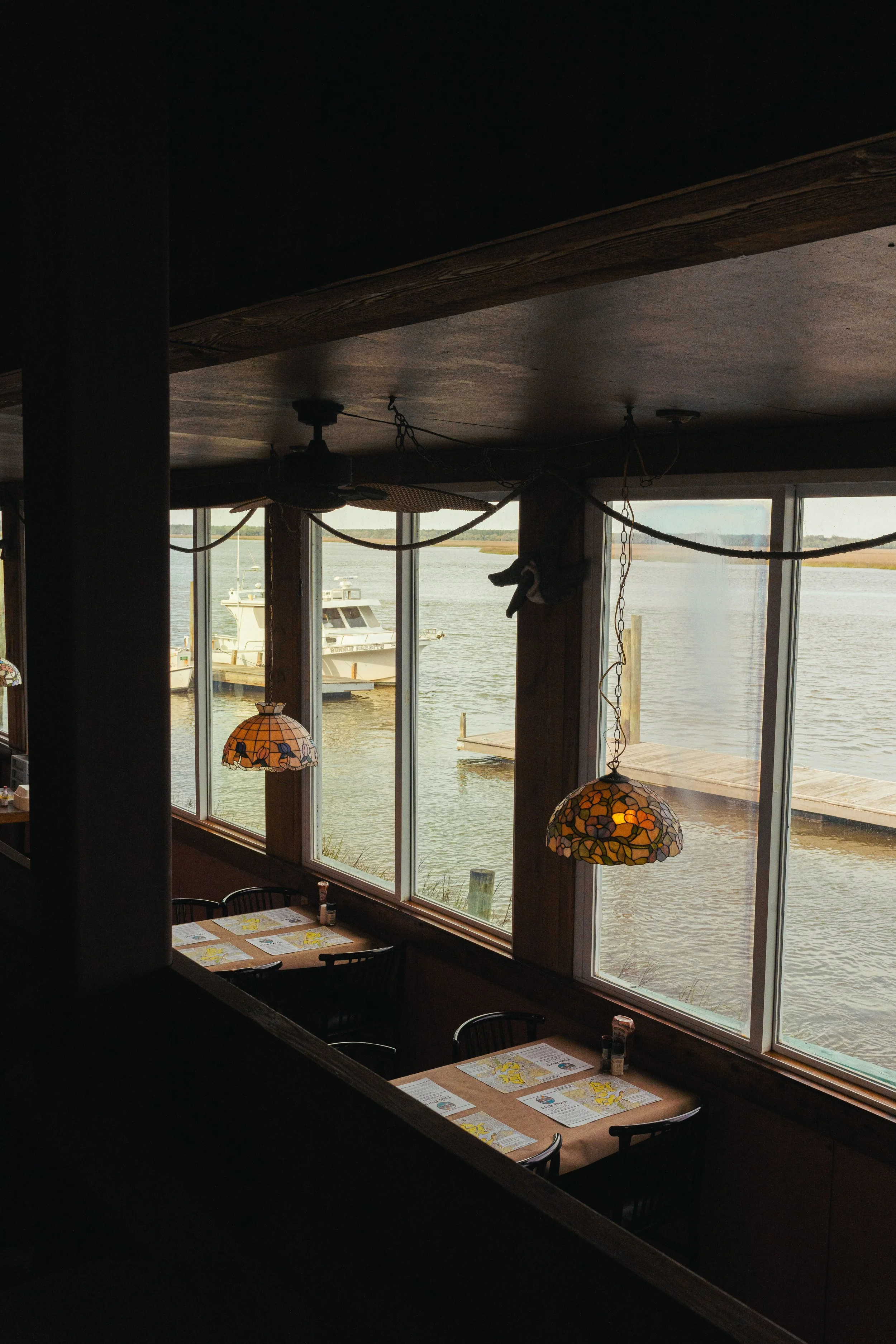Interior view of a lakeside restaurant with large windows showing boats and water outside. Hanging stained glass lamps provide lighting over tables with menus and condiments.