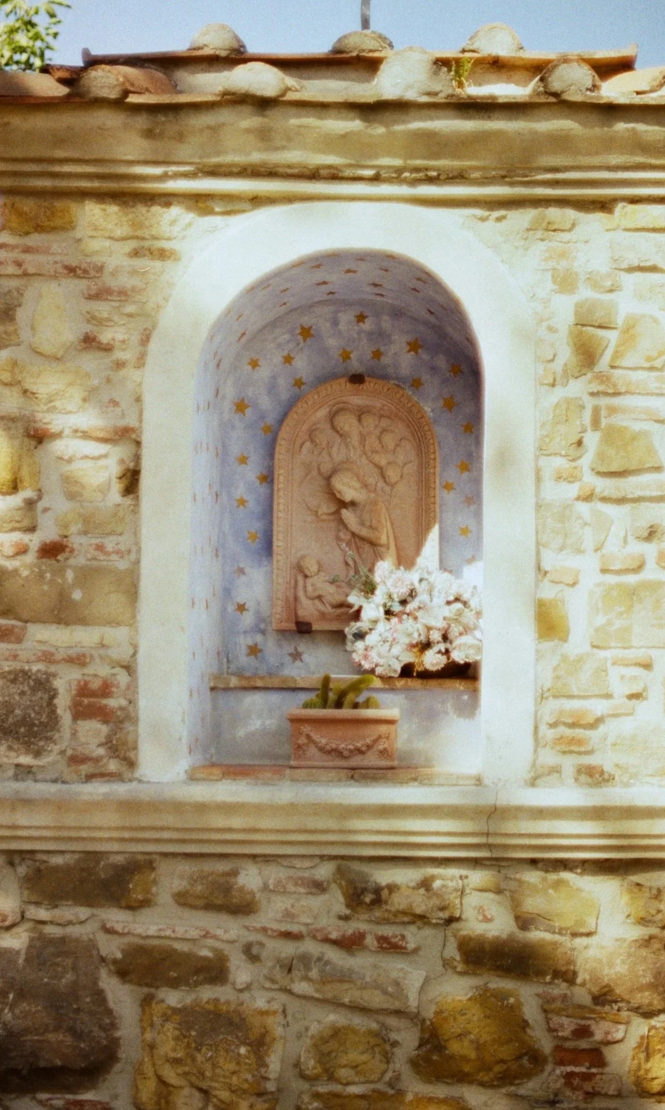 A religious shrine embedded in a stone brick wall, featuring a carved depiction of a biblical scene, with a bouquet of white flowers and a potted cactus placed below.