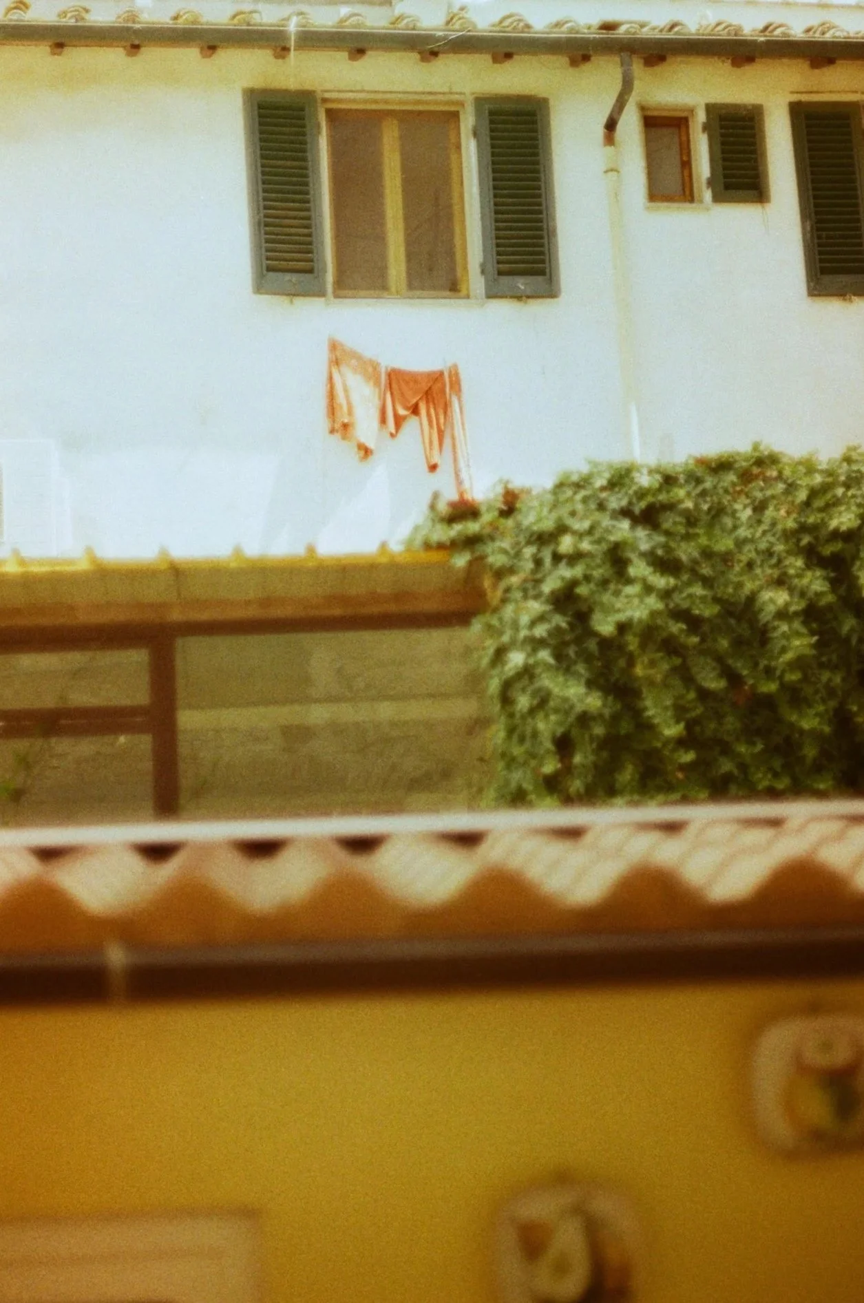 An exterior view of a house wall with three windows, two with open shutters. There are clothes hanging outside the window in the middle. A bush partially obscures the bottom right corner. A wooden bench is visible at the bottom of the image.