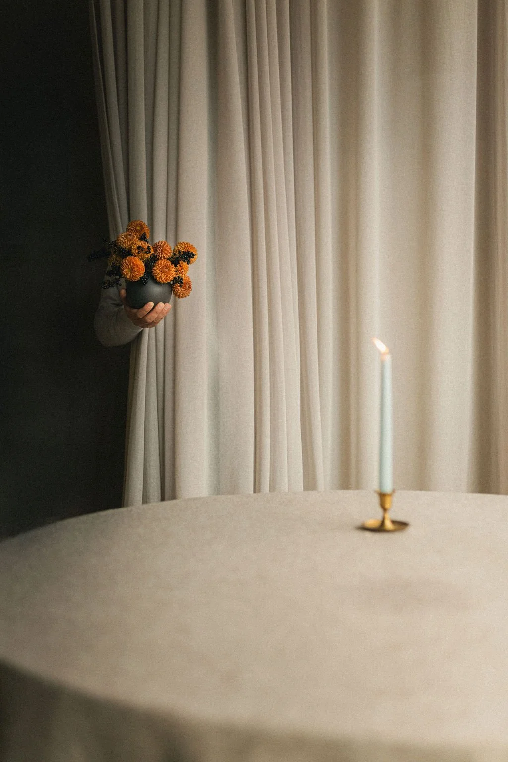 Hand holding a dahlia posy from behind a linen curtain beside a candlelit table, editorial floral photography Montréal for Art et Fleurs