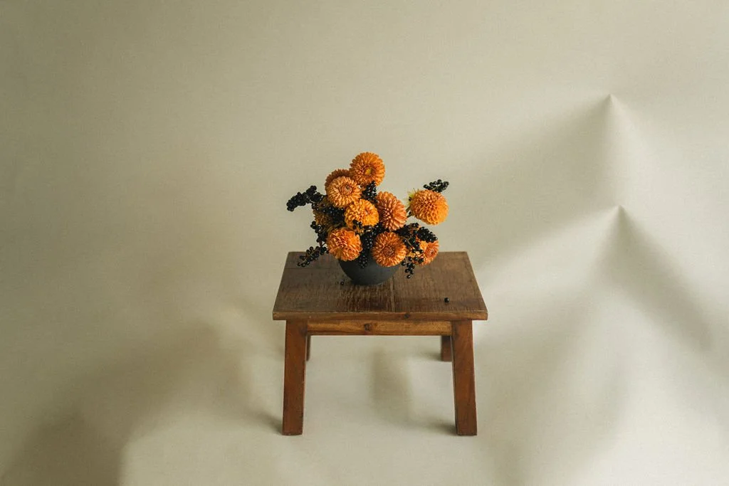 Orange dahlias and dark berries in a black vase on a wooden stool — floral photography in Montréal by A. Wood for Art et Fleurs