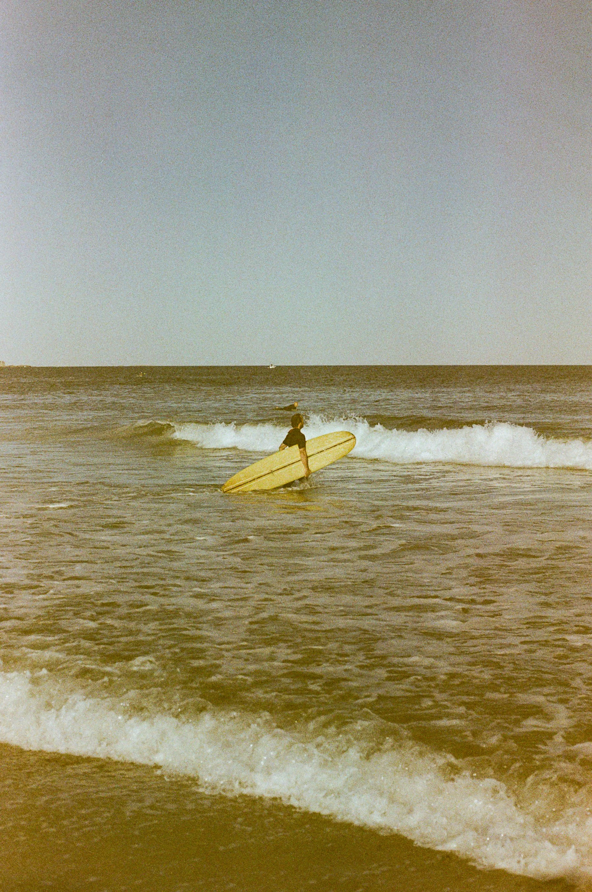 A person holding a surfboard while standing in the ocean near the shore, with waves and a distant boat visible.