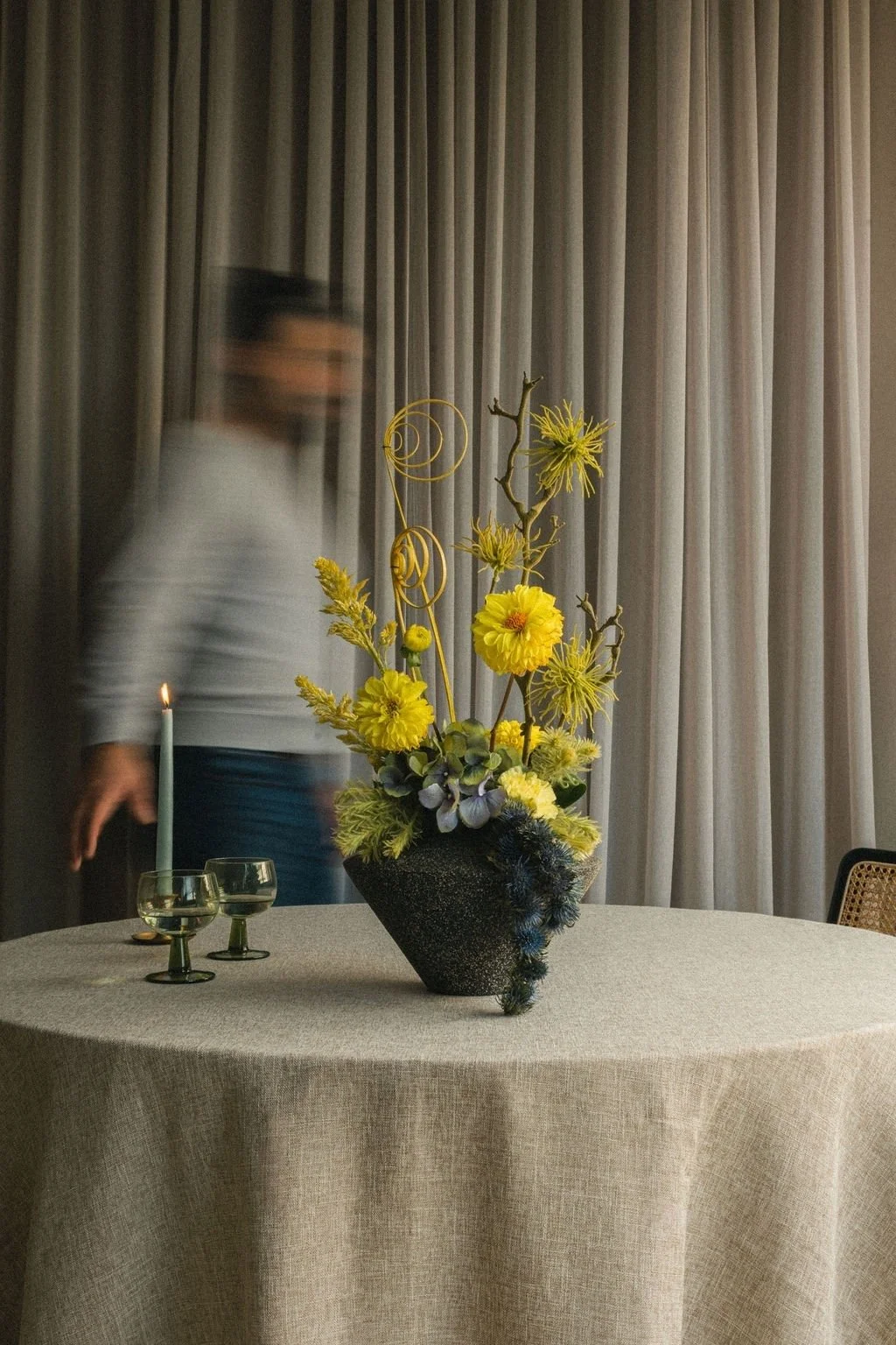 A blurred figure passing behind yellow chrysanthemums on a linen-draped table, fall floral photography for Art et Fleurs, Montréal