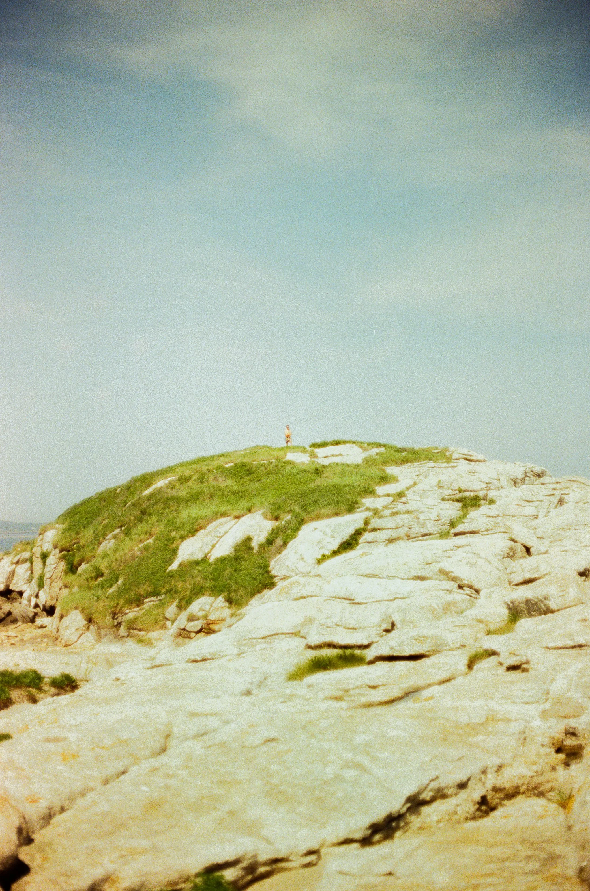 A small person standing on top of a grassy hill near white rocky cliffs under a partly cloudy sky.