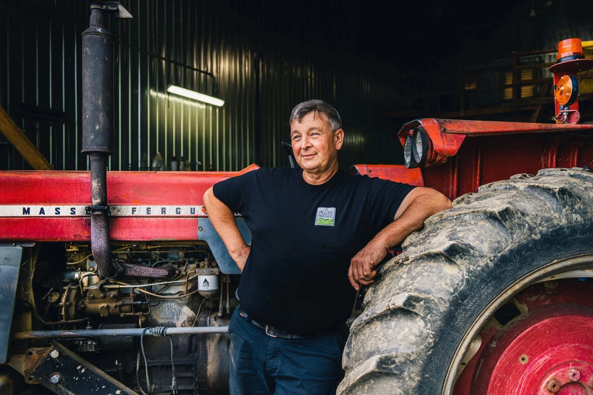A farmer leaning against a red tractor in a barn, agricultural portrait photography by A. Wood