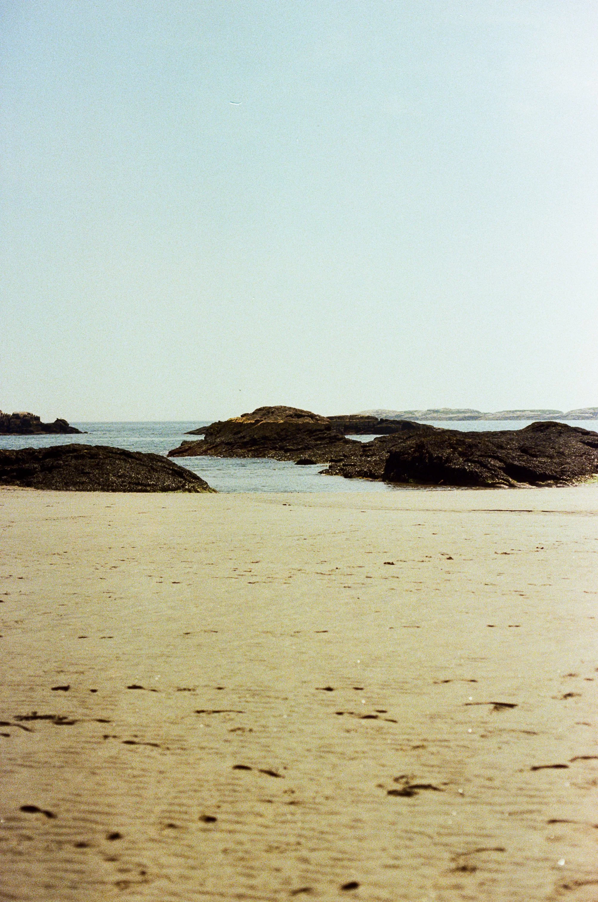 Sandy beach with rocks and calm ocean water, distant landforms, and a hazy sky with a faint airplane visible overhead.
