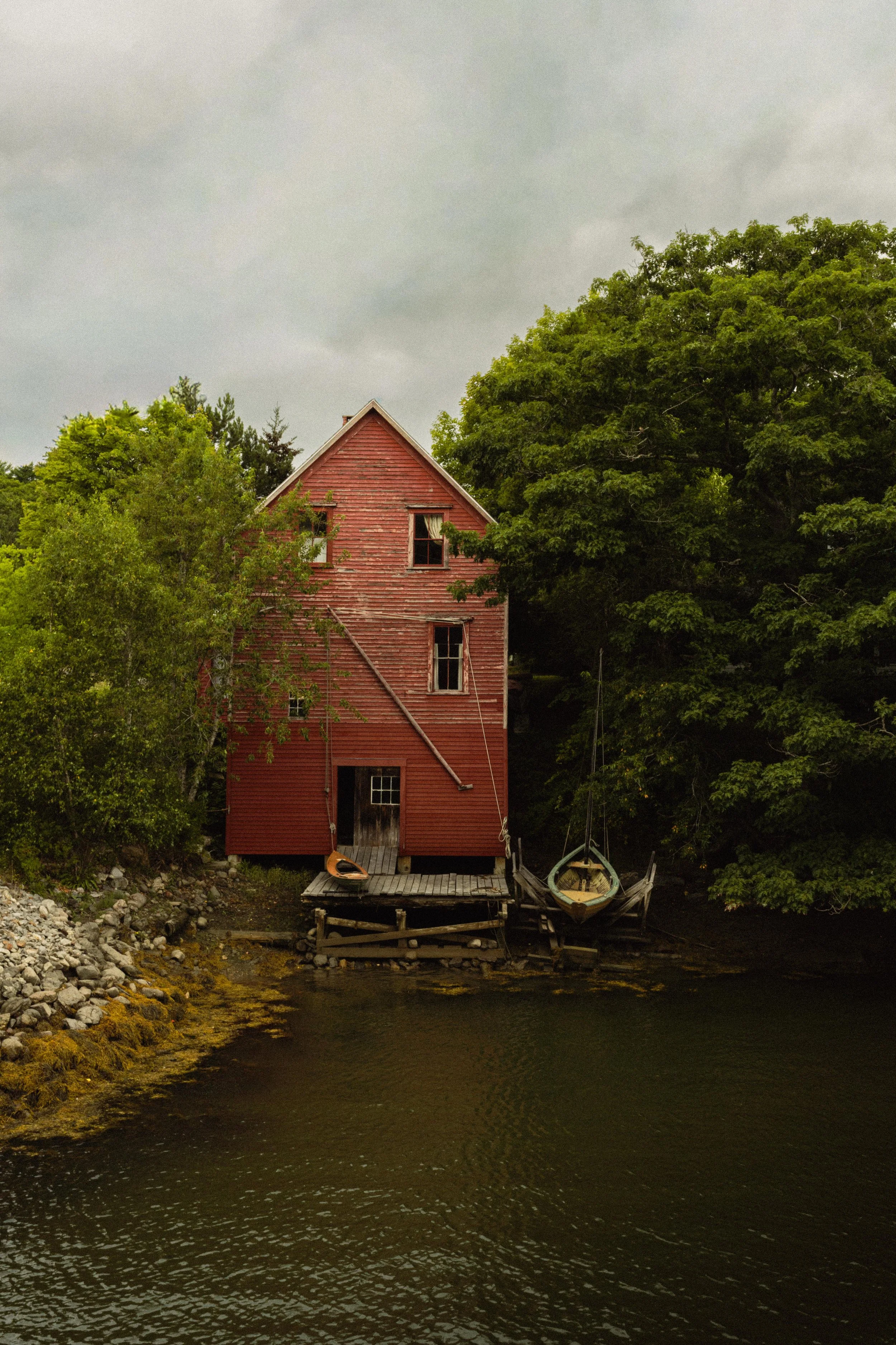 A red wooden house with boats docked at its base, surrounded by green trees, beside a body of water under an overcast sky.