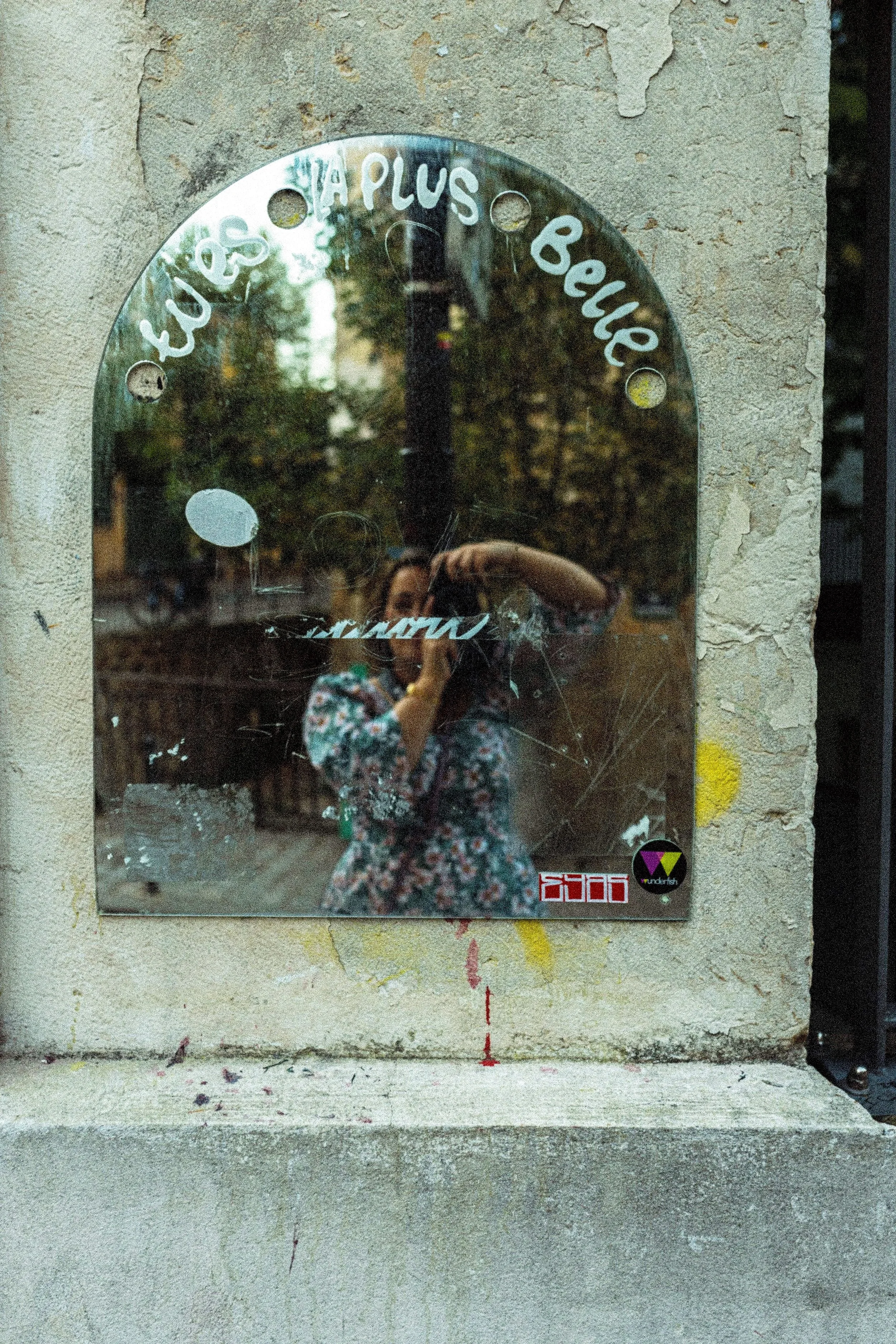 A small mirror attached to a textured wall, reflecting a woman taking a photo with a camera, wearing a floral dress, with trees and street visible in the reflection.