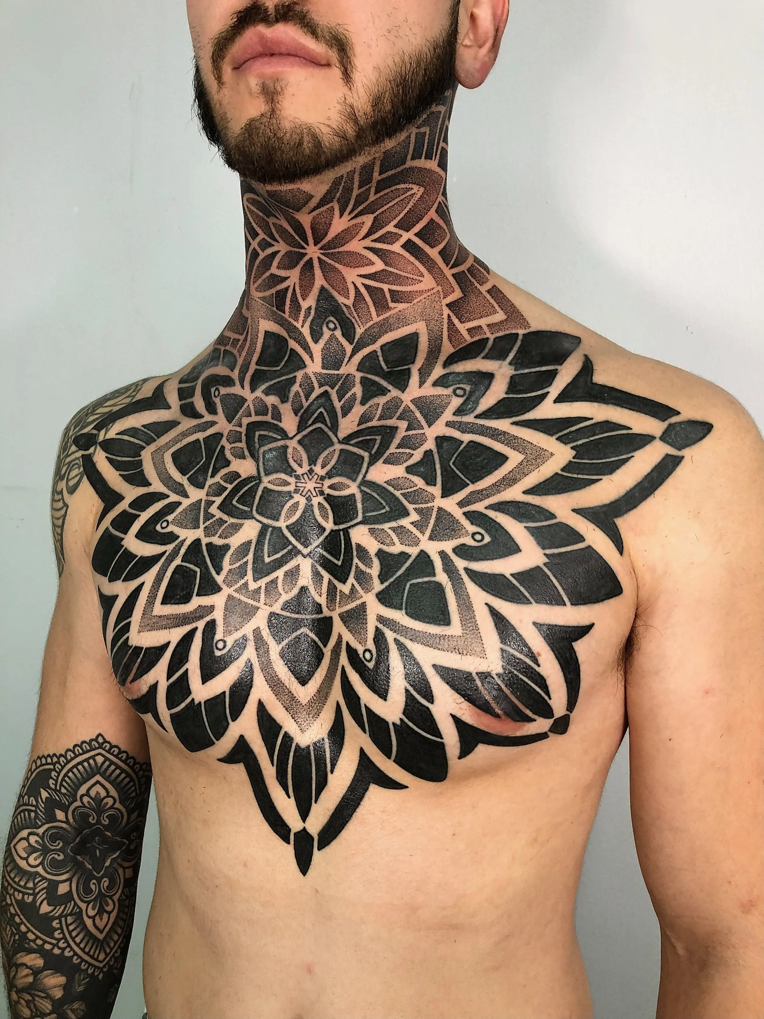 A shirtless man with multiple black geometric and floral tattoos covering his chest, neck, and arm.