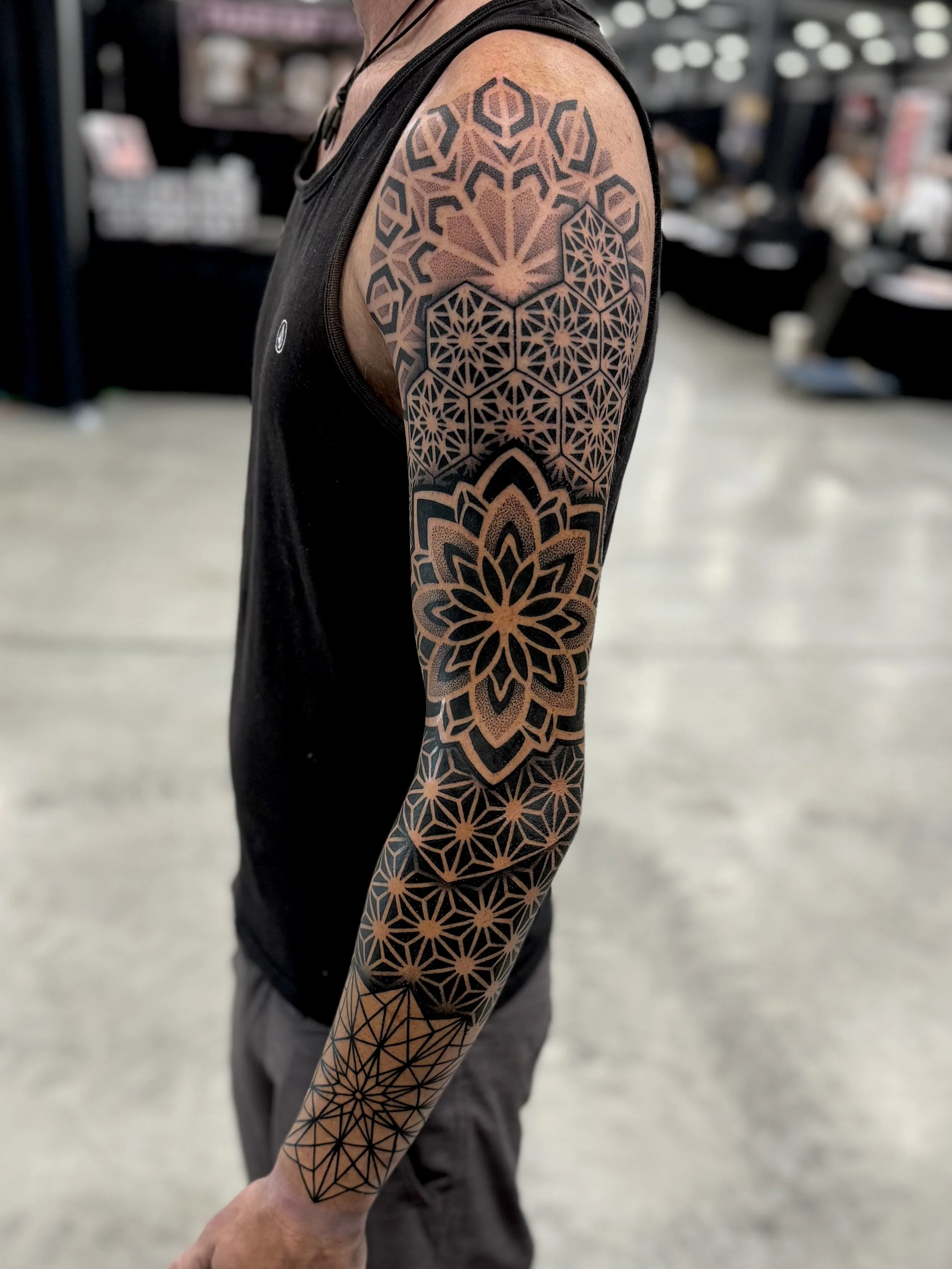 A person's arm with geometric black and tan tattoo designs extending from the shoulder to the wrist, wearing a sleeveless black shirt in a public indoor space.