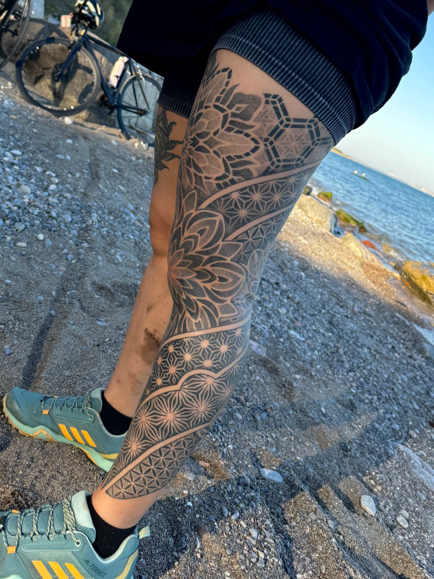 Close-up of a person's tattooed leg, wearing running shoes, on a rocky beach near the water, with a bicycle in the background.