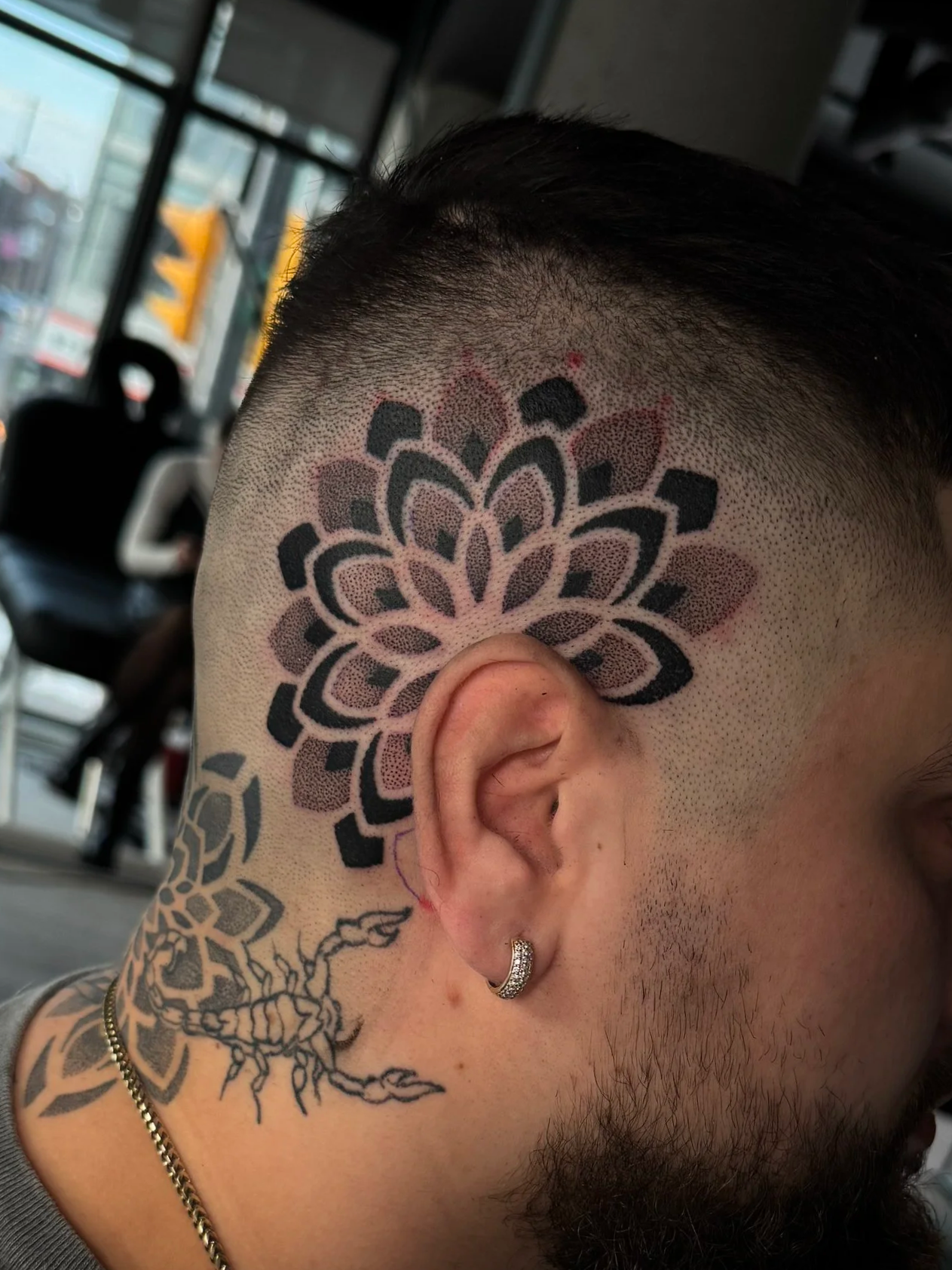 Close-up of a man's head with a geometric floral tattoo on his scalp and a smaller tattoo of a scorpion on his neck. He has a short, dark haircut and wears a silver hoop earring.