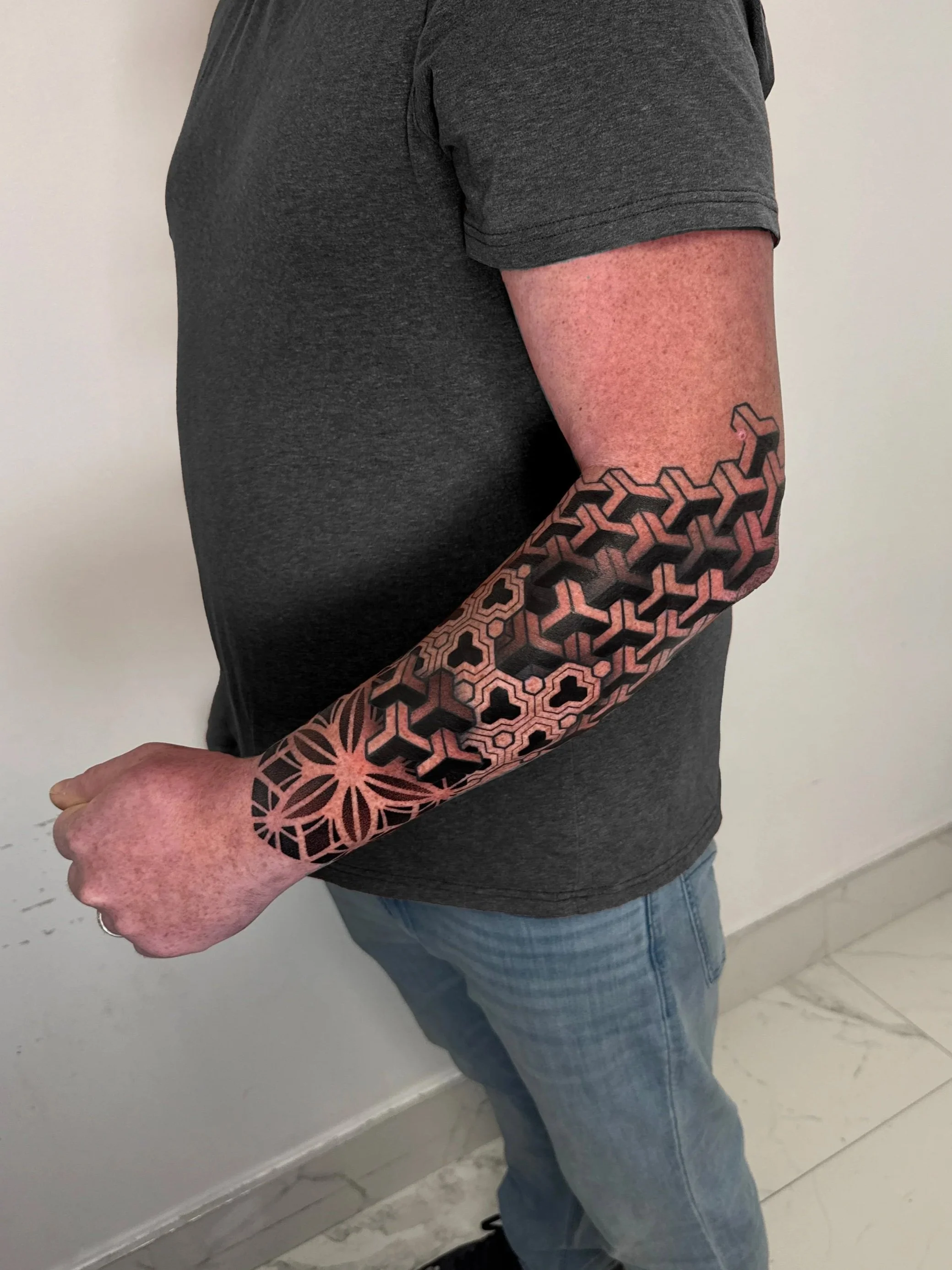 A person wearing a dark grey T-shirt and blue jeans, showing a sleeve tattoo with geometric patterns on their right arm.