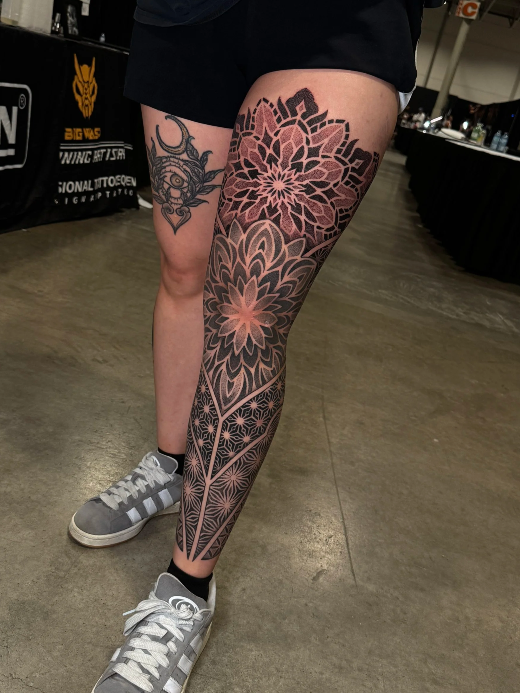 Person standing indoors wearing gray sneakers and black shorts with extensive geometric floral tattoo designs on their legs, which are crossed at the ankles.