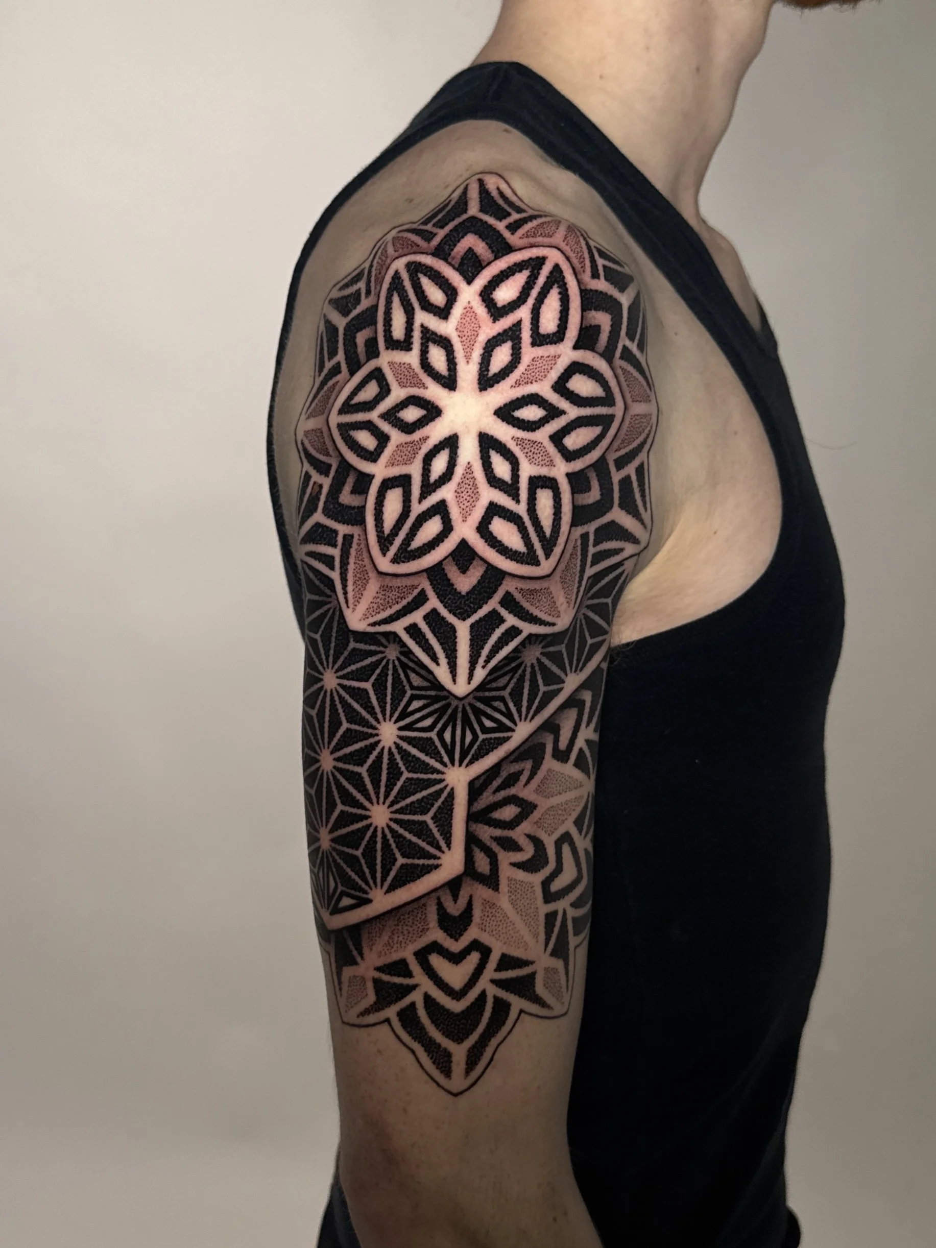 A person's upper arm and shoulder featuring a large, intricate geometric tattoo design in black and light pink ink.