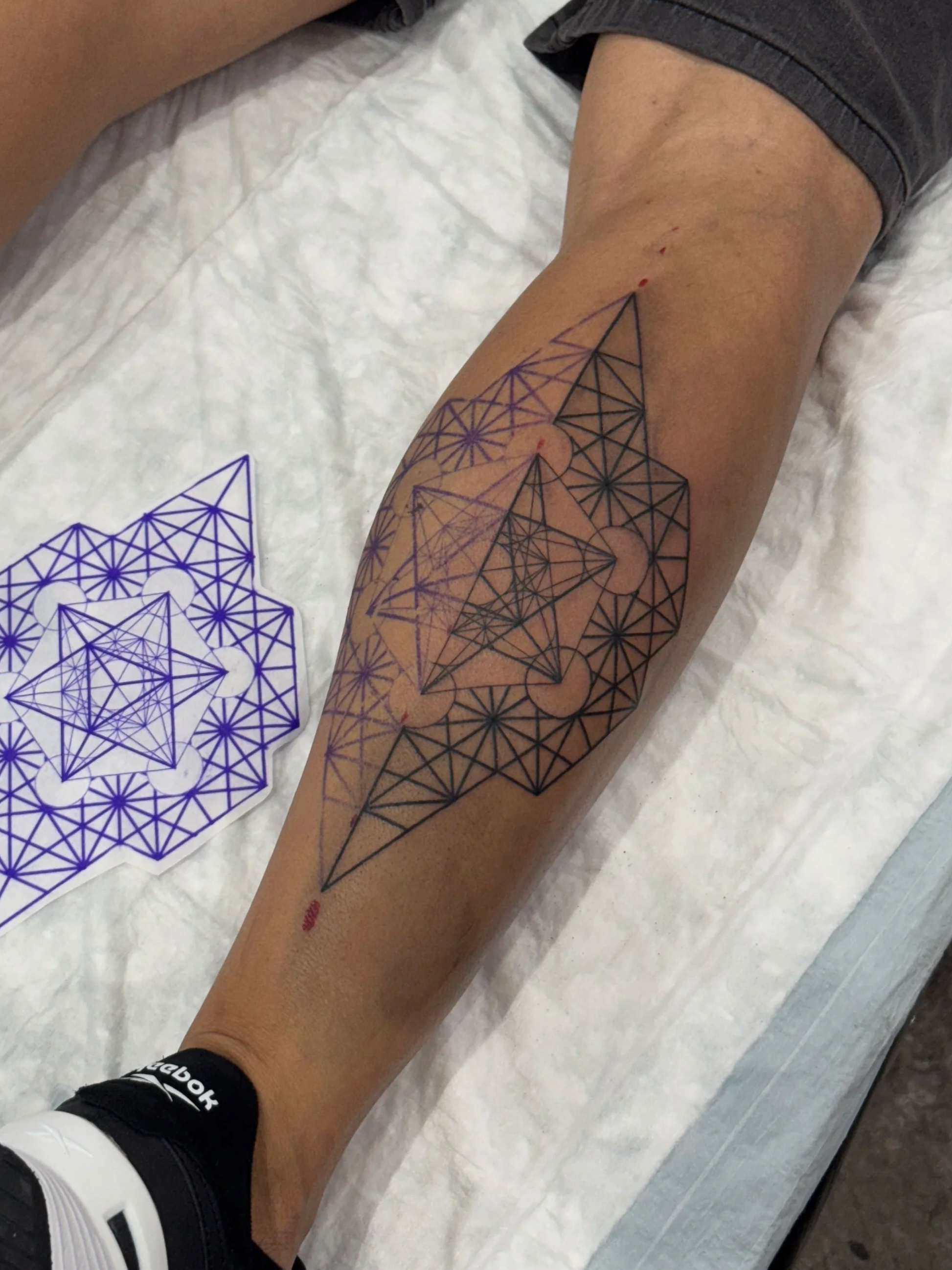 A leg with a geometric tattoo covering the thigh, sitting on a white sheet with tattoo stencil paper nearby.