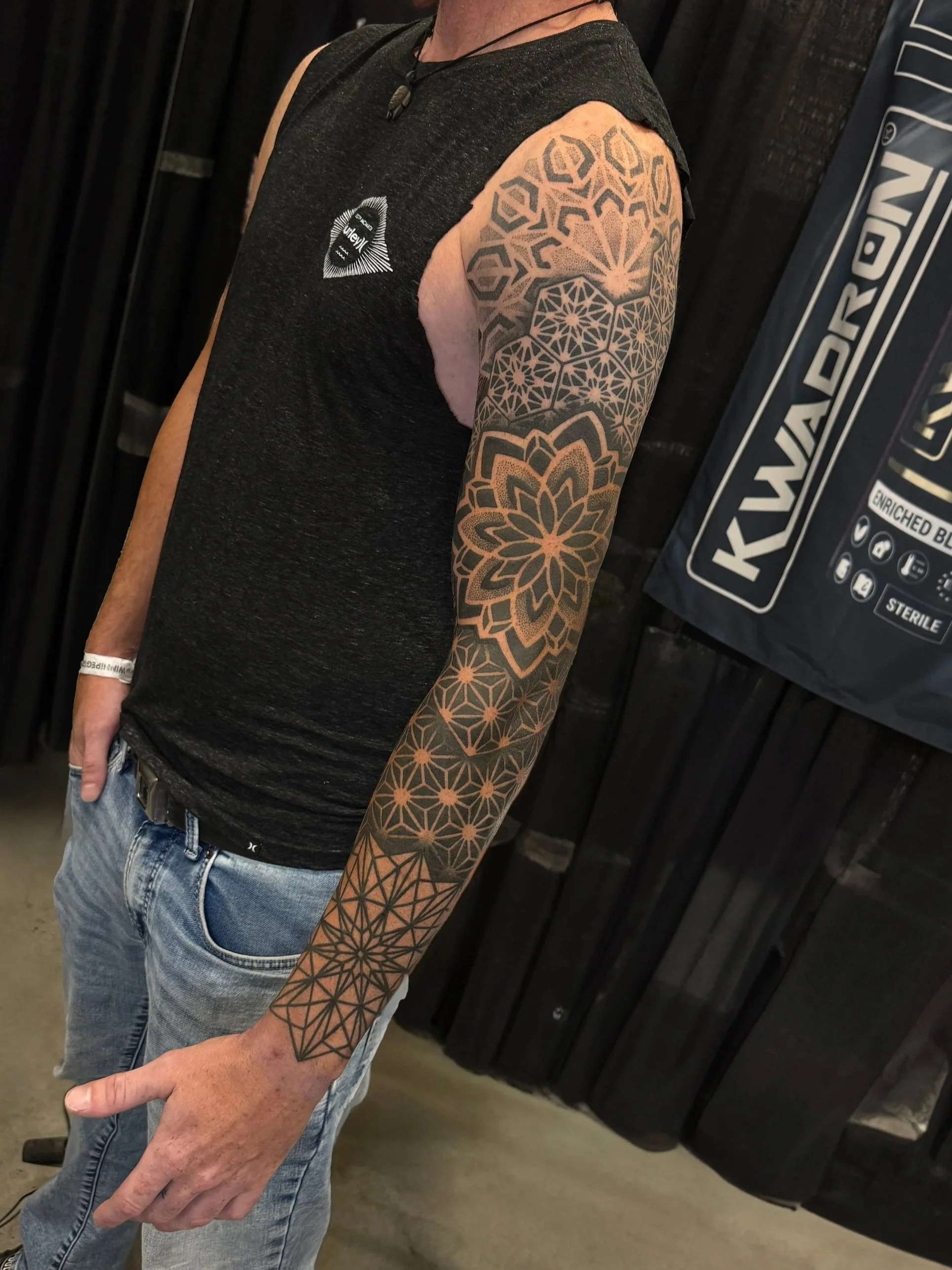 Close-up of a person with a geometric pattern tattoo on their arm, wearing a black sleeveless shirt with a logo, and jeans, standing indoors with a black curtain or wall behind them.
