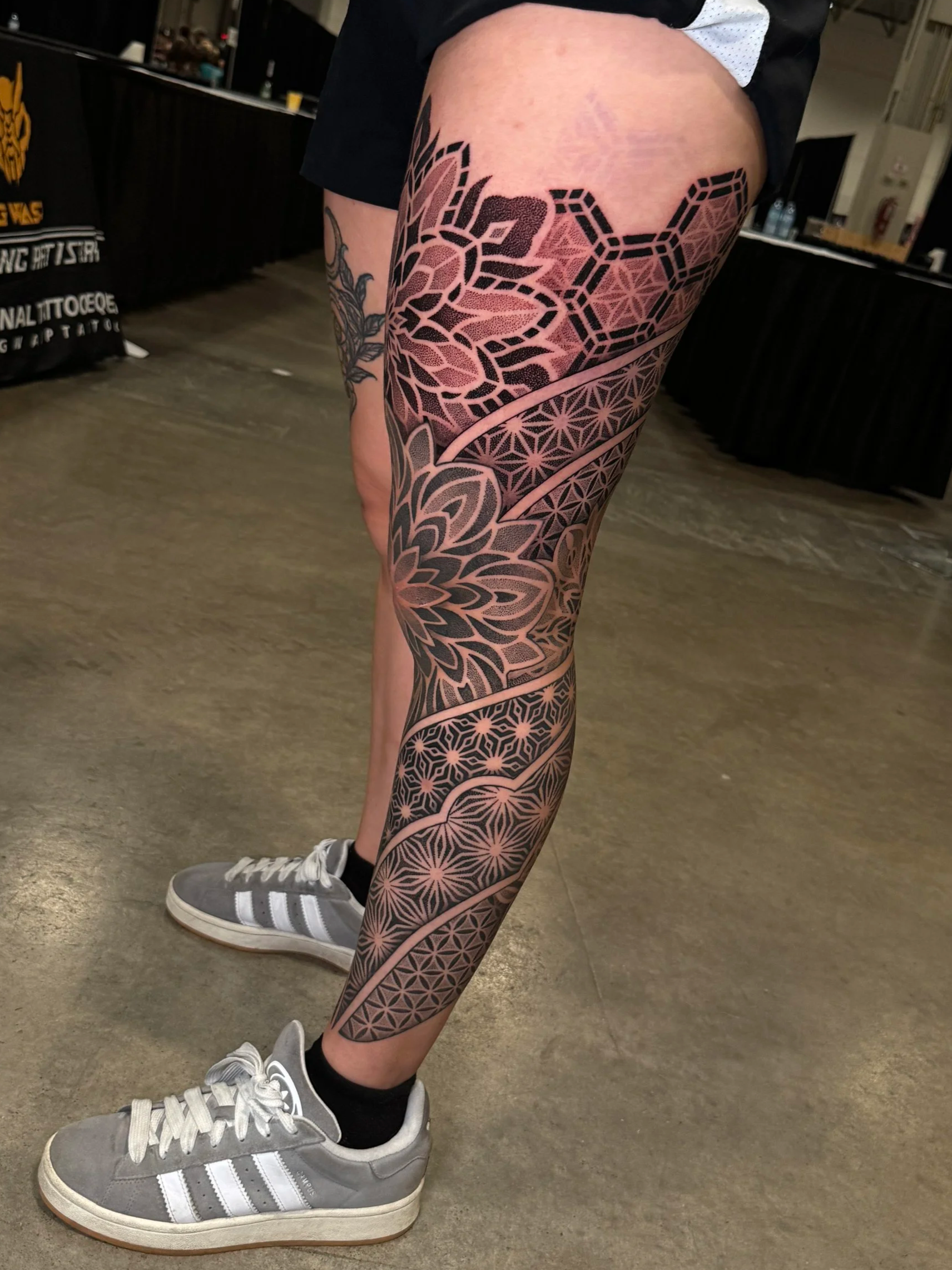 Close-up of a person's leg showcasing a detailed geometric tattoo extending from the thigh to the ankle, with the person wearing gray Adidas sneakers and black socks, standing on a concrete floor in an indoor setting.