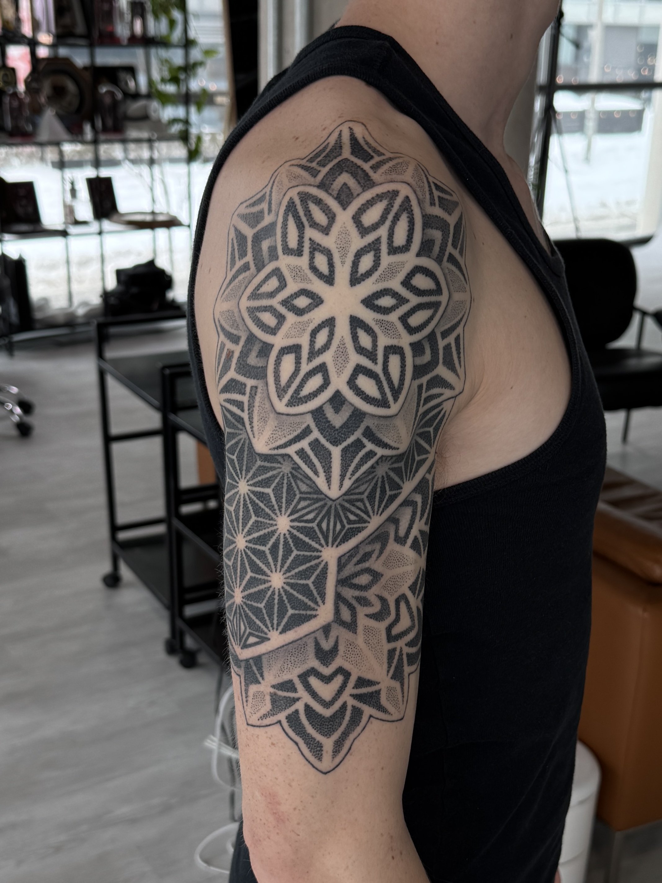 A person's upper arm and shoulder with a black geometric tattoo on the shoulder extending down the arm, featuring intricate patterns and shapes.