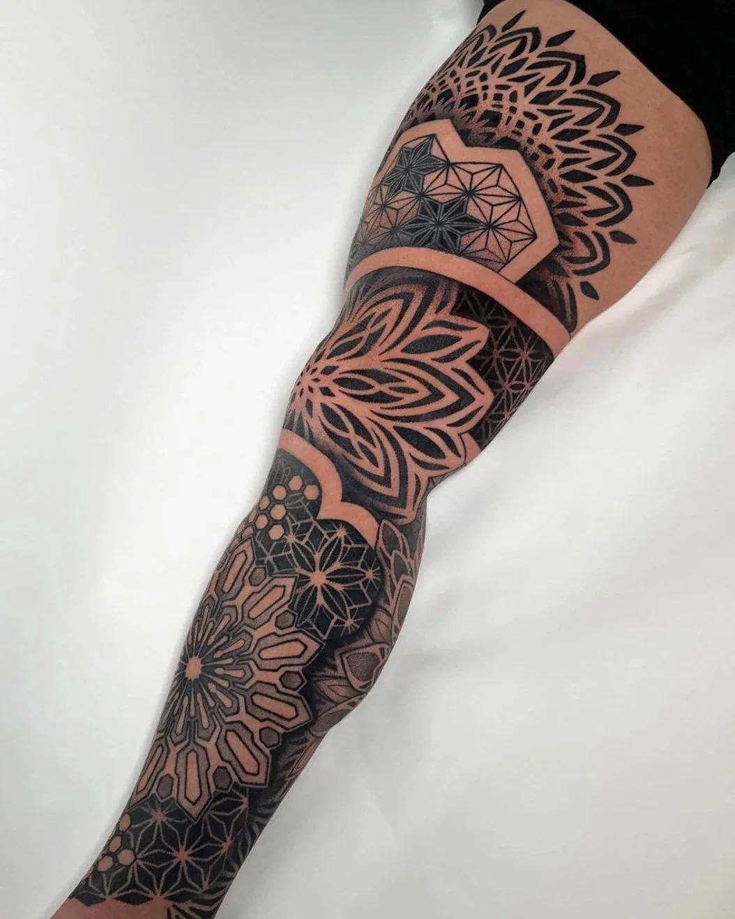A person's arm with a full sleeve tattoo featuring geometric and floral patterns in black and skin tone shading.