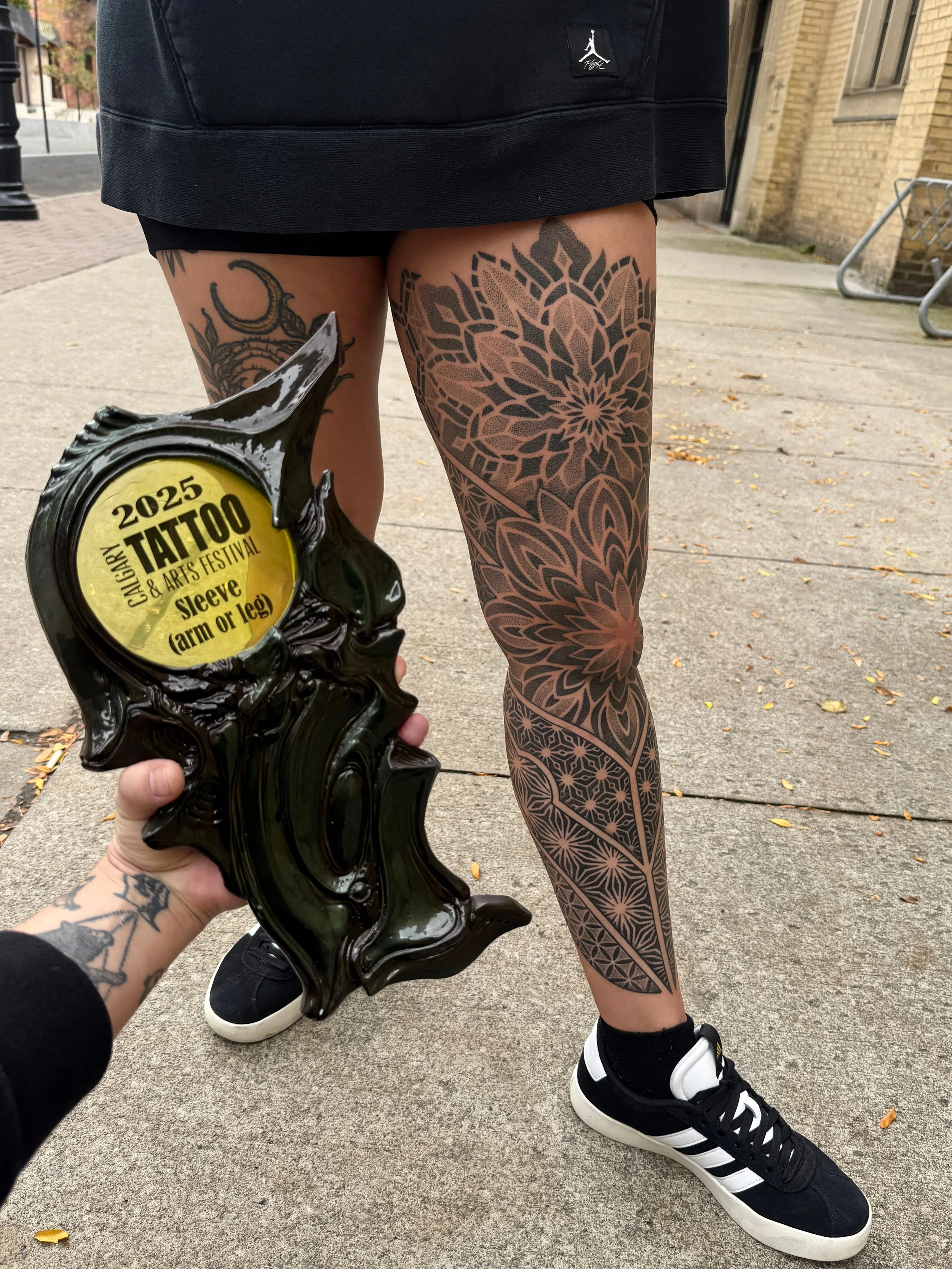 Close-up of a woman with detailed black tattoos on her leg, standing on a sidewalk. She wears black and white sneakers, black socks, and a short black skirt. A person's hand holds a black sculpture with a yellow sticker reading '2025 Tattoo & Arts Festival Sleave (arm or leg)' in front of her.
