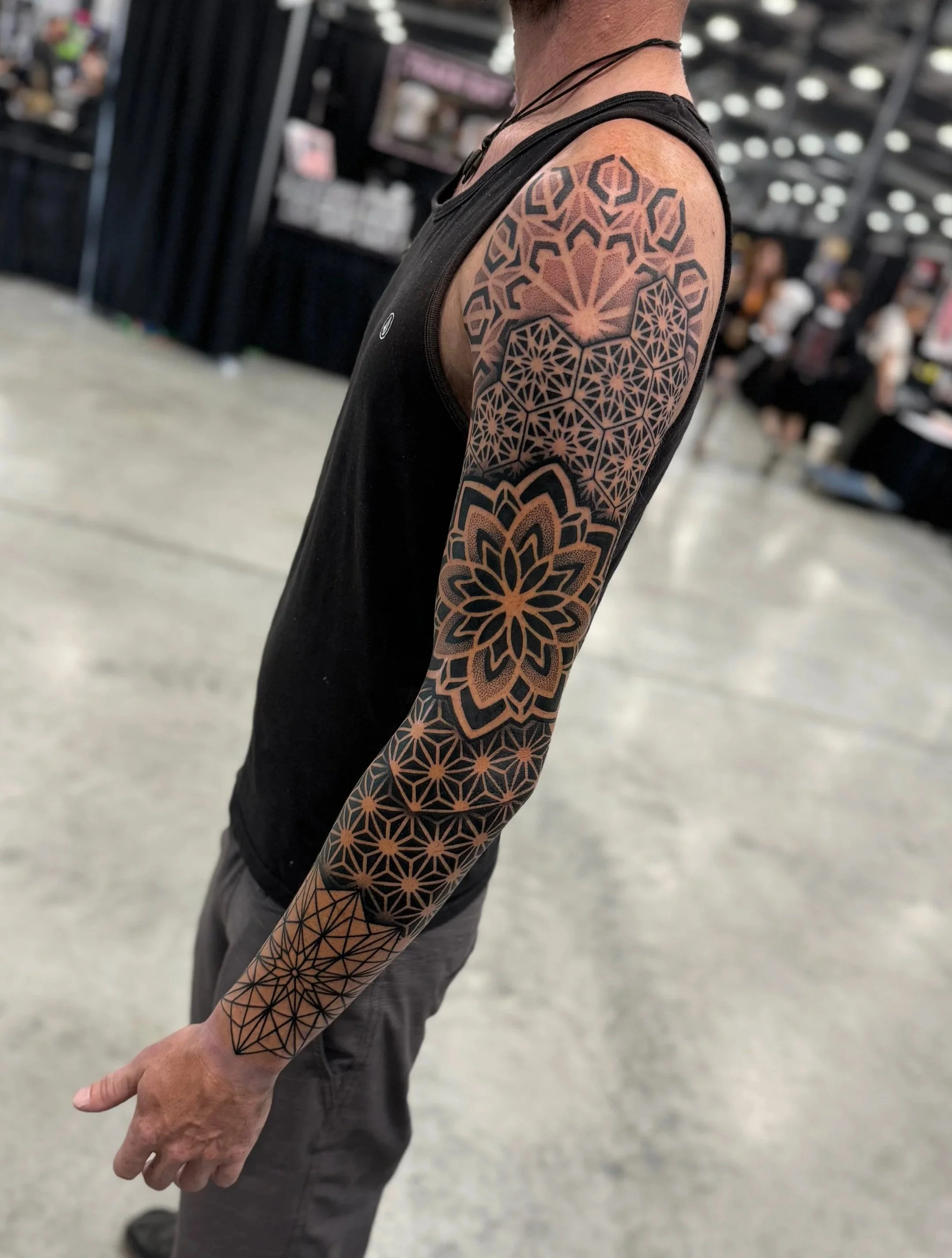 A person with a geometric tattoo sleeve on their arm, standing in an indoor event or convention, wearing a black sleeveless shirt.