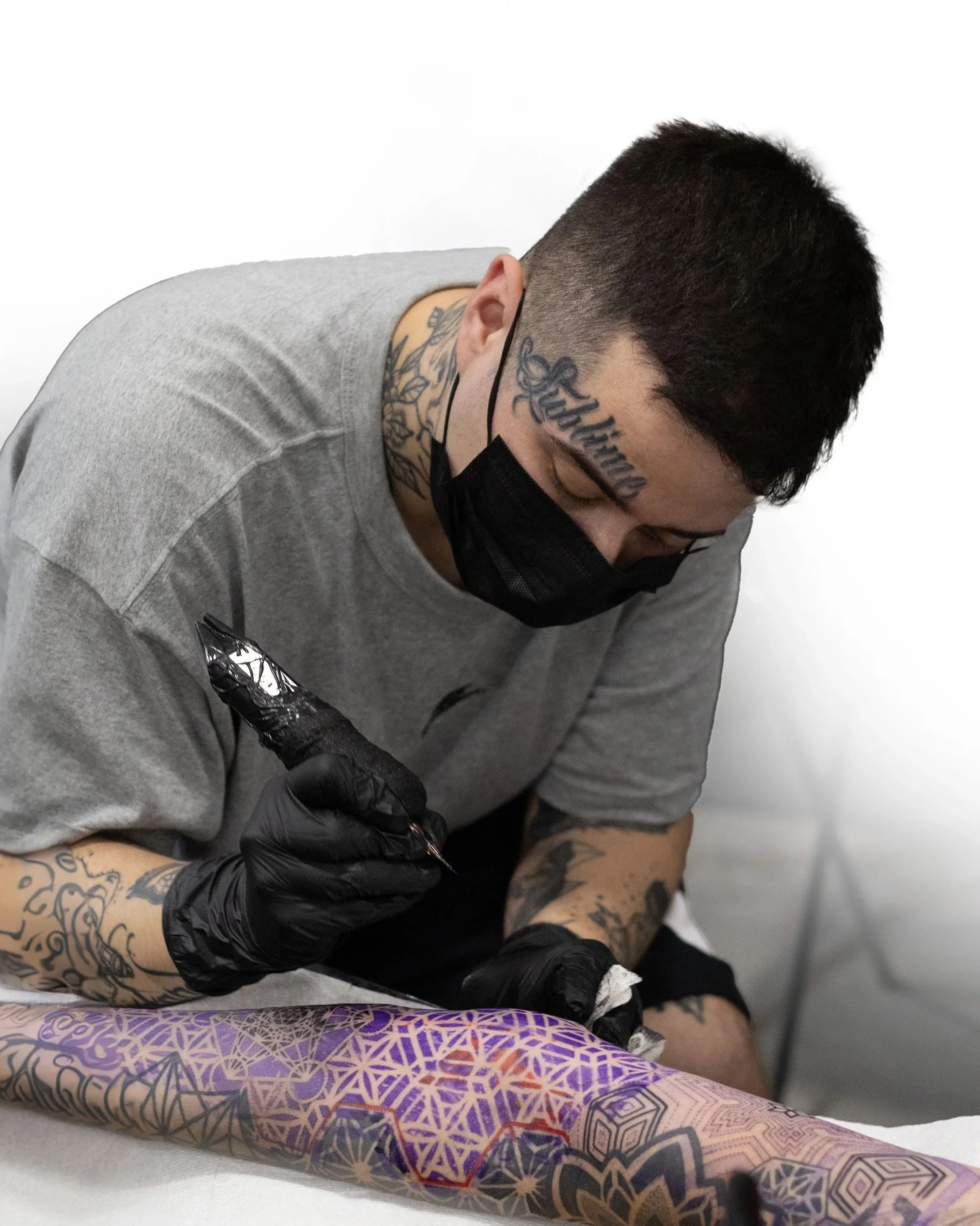 A person with tattoos getting a tattoo on their arm by a tattoo artist wearing black gloves. The person's head has a tattoo that says "Sutherland." The person's arm has a colorful geometric tattoo.