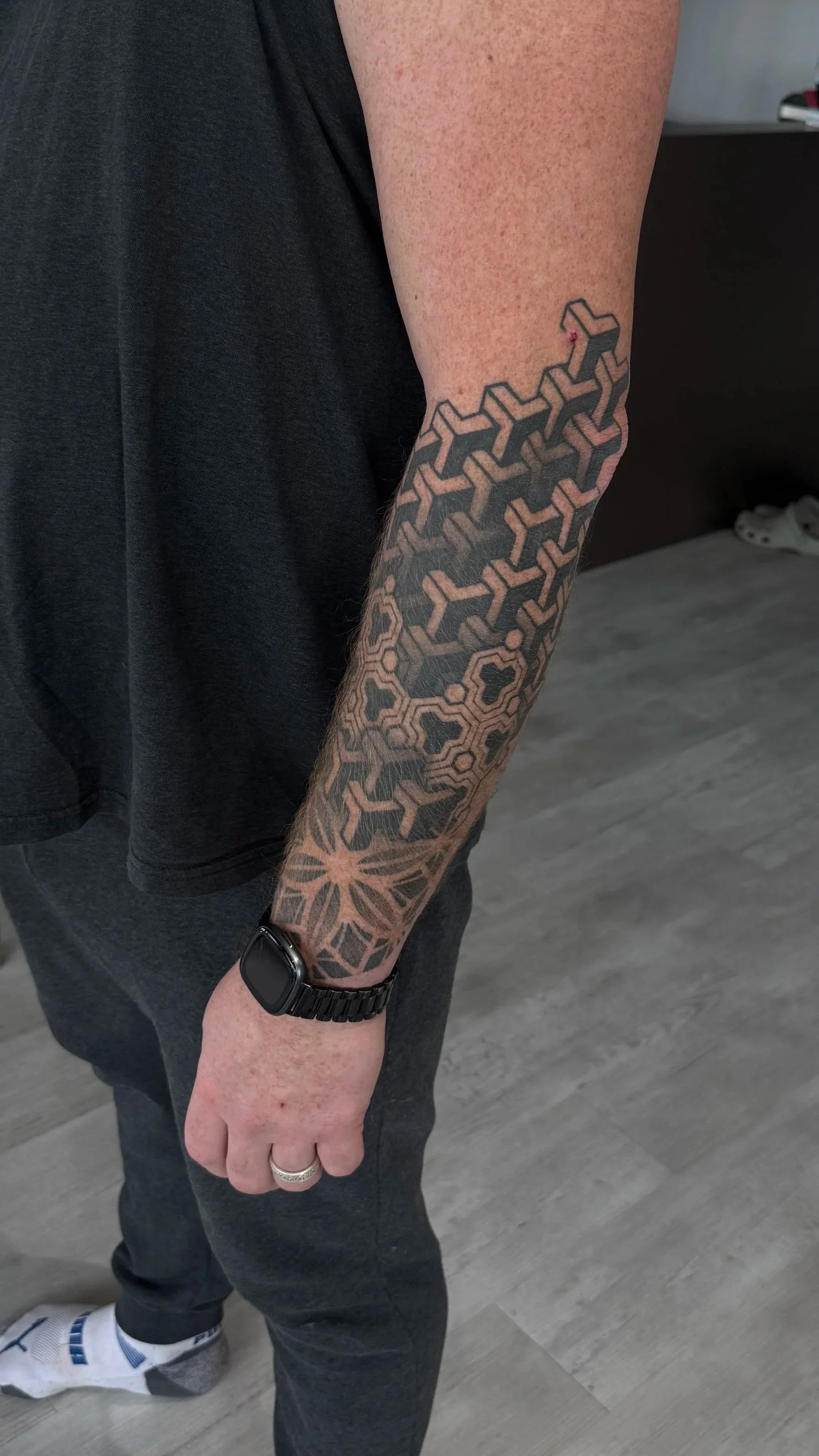 A person's arm with a large geometric tattoo extending from the wrist to the elbow, featuring an intricate pattern of interlocking shapes. The person is wearing a black wristwatch and a silver ring on their finger, dressed in dark pants and a black shirt, with a grey floor and a dark piece of furniture in the background.
