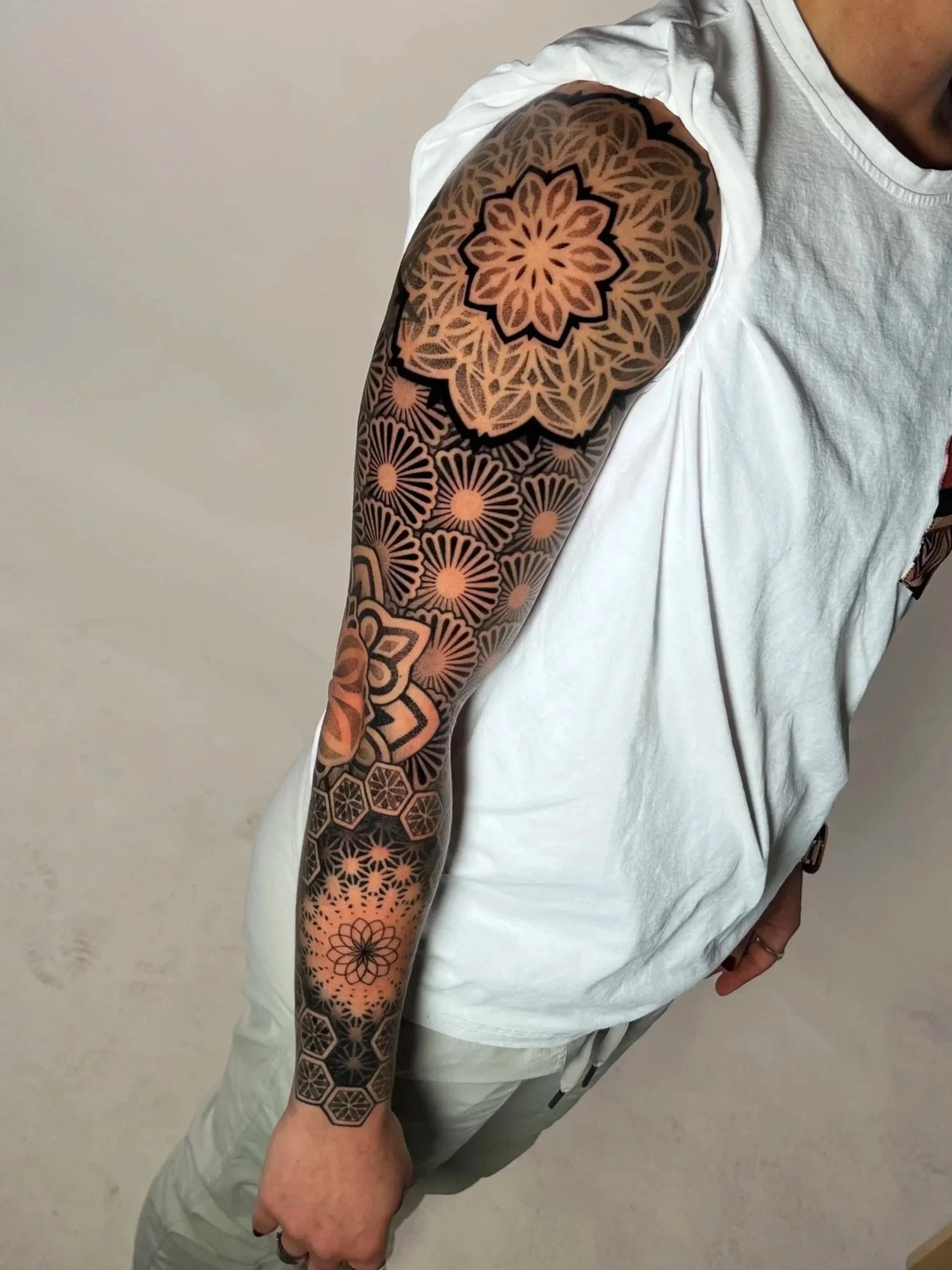 A person wearing a white t-shirt and grey pants shows a detailed geometric tattoo sleeve on their left arm, featuring various patterns including a large flower design and hexagonal shapes.