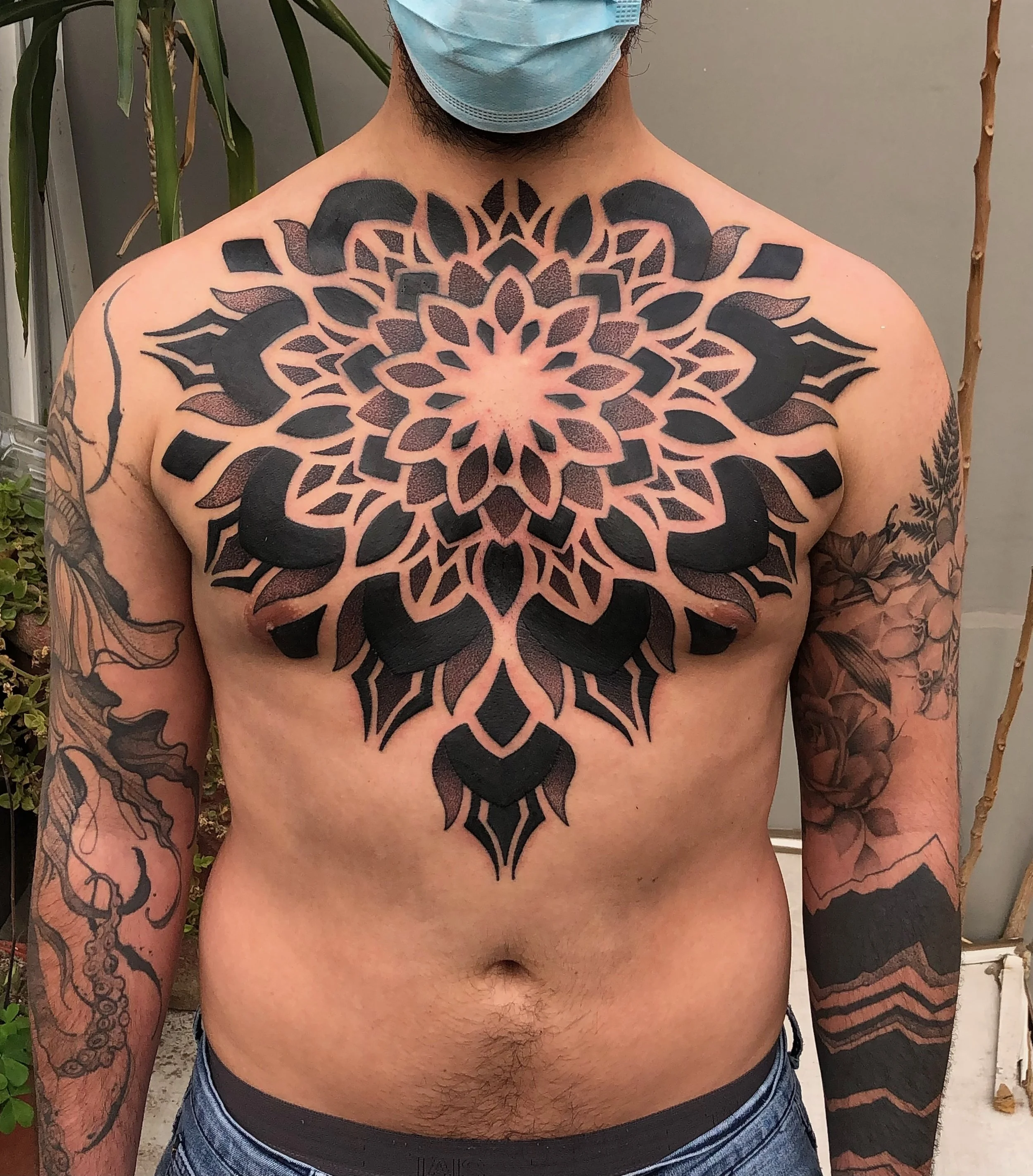 A shirtless man with multiple tattoos, including a large symmetrical black and gray geometric mandala on his chest extending to his shoulders. He wears a blue surgical mask, and has tattoos on his arms of floral and abstract designs. He is standing in front of a gray wall with plants in the background.