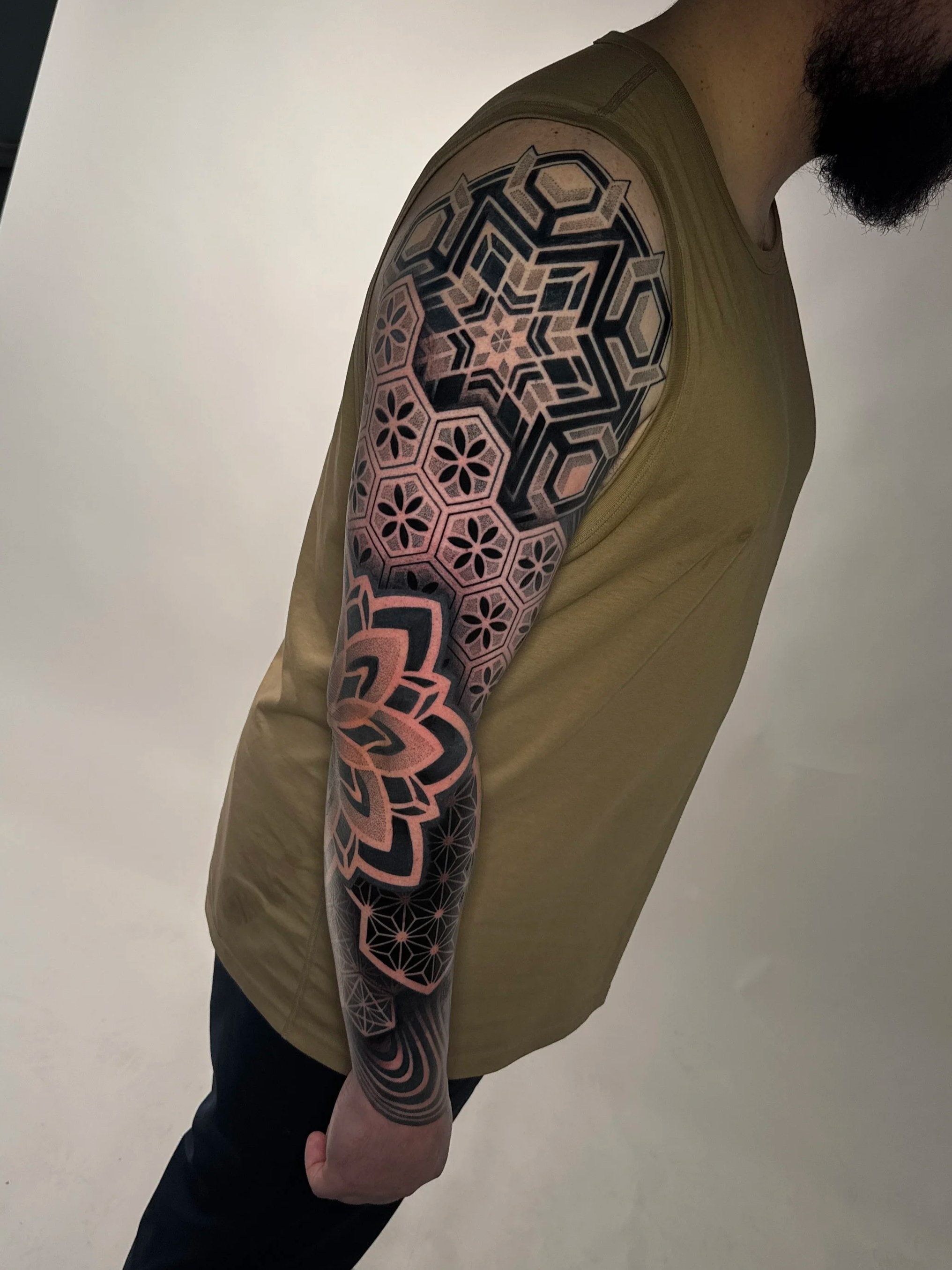 Close-up of a person's arm with a large geometric tattoo featuring black and pink patterns, wearing a beige sleeveless shirt.