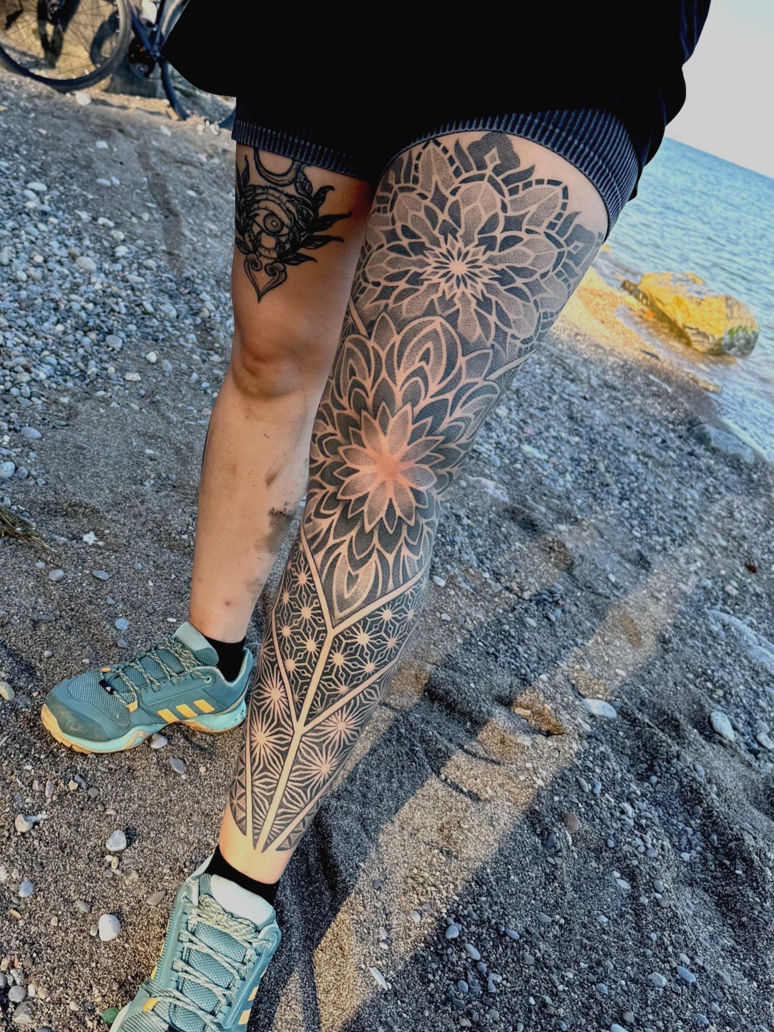 Close-up of a person wearing teal and yellow running shoes, standing on rocky beach with tattoos on their legs, near water.