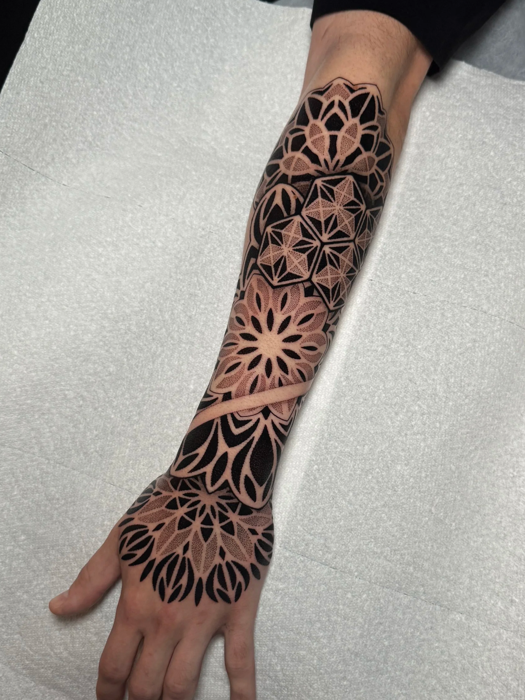 A person's arm with a geometric tattoo design covering the wrist to the upper forearm, featuring black and flesh-colored patterns resembling mandalas or floral motifs.
