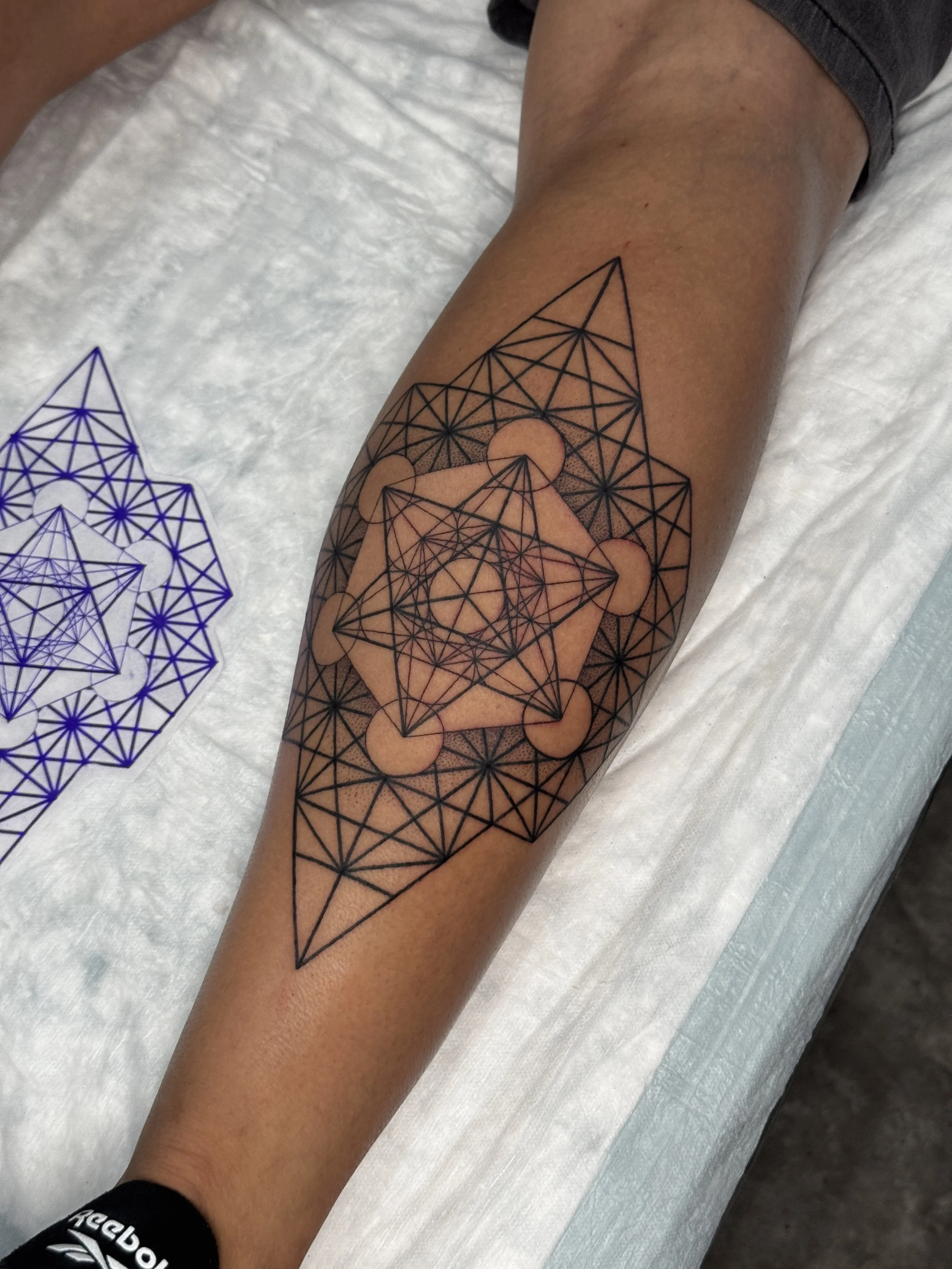A person's arm with a geometric tattoo featuring interconnected lines and circles, with a paper stencil of the tattoo design nearby.