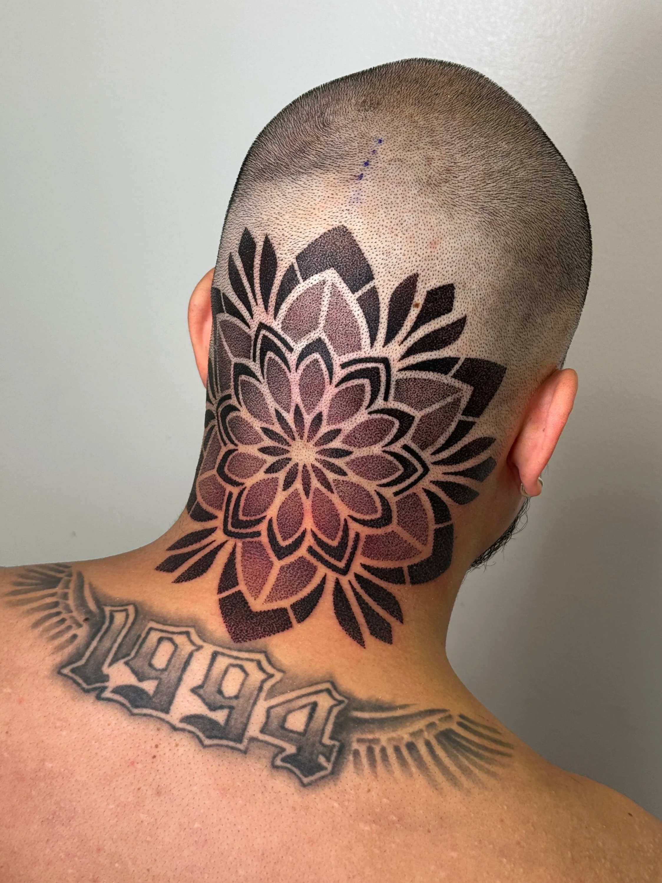 Close-up of a person's upper arm, shoulder, and neck, with a floral patterned tattoo covering the shoulder and extending down the arm. The person has a shaved head with small dots marked with blue ink, possibly for a tattoo or piercing procedure.