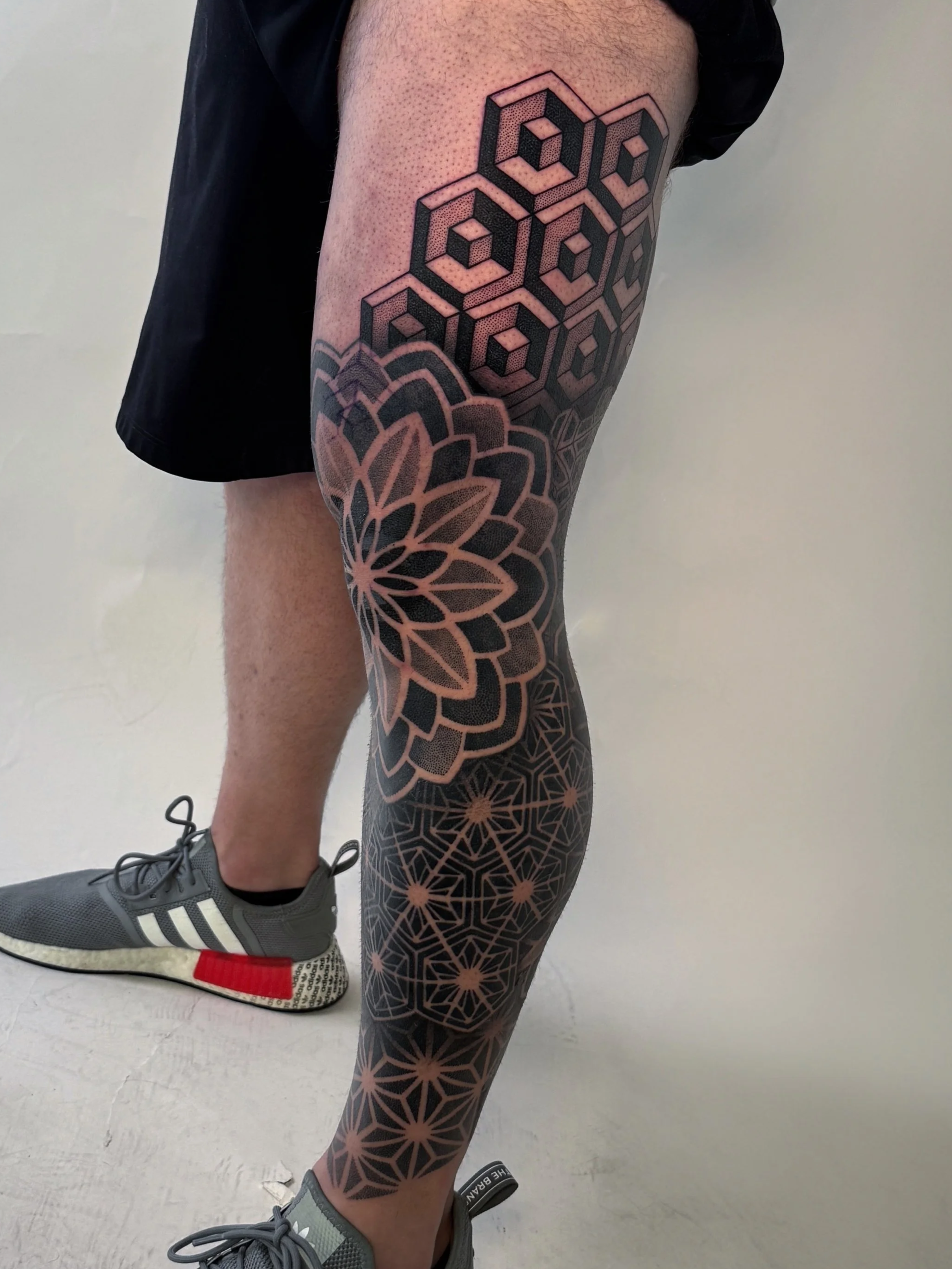 Tattooed leg with geometric and floral black ink designs, including overlapping cubes and a large flower pattern, wearing gray athletic shoes
